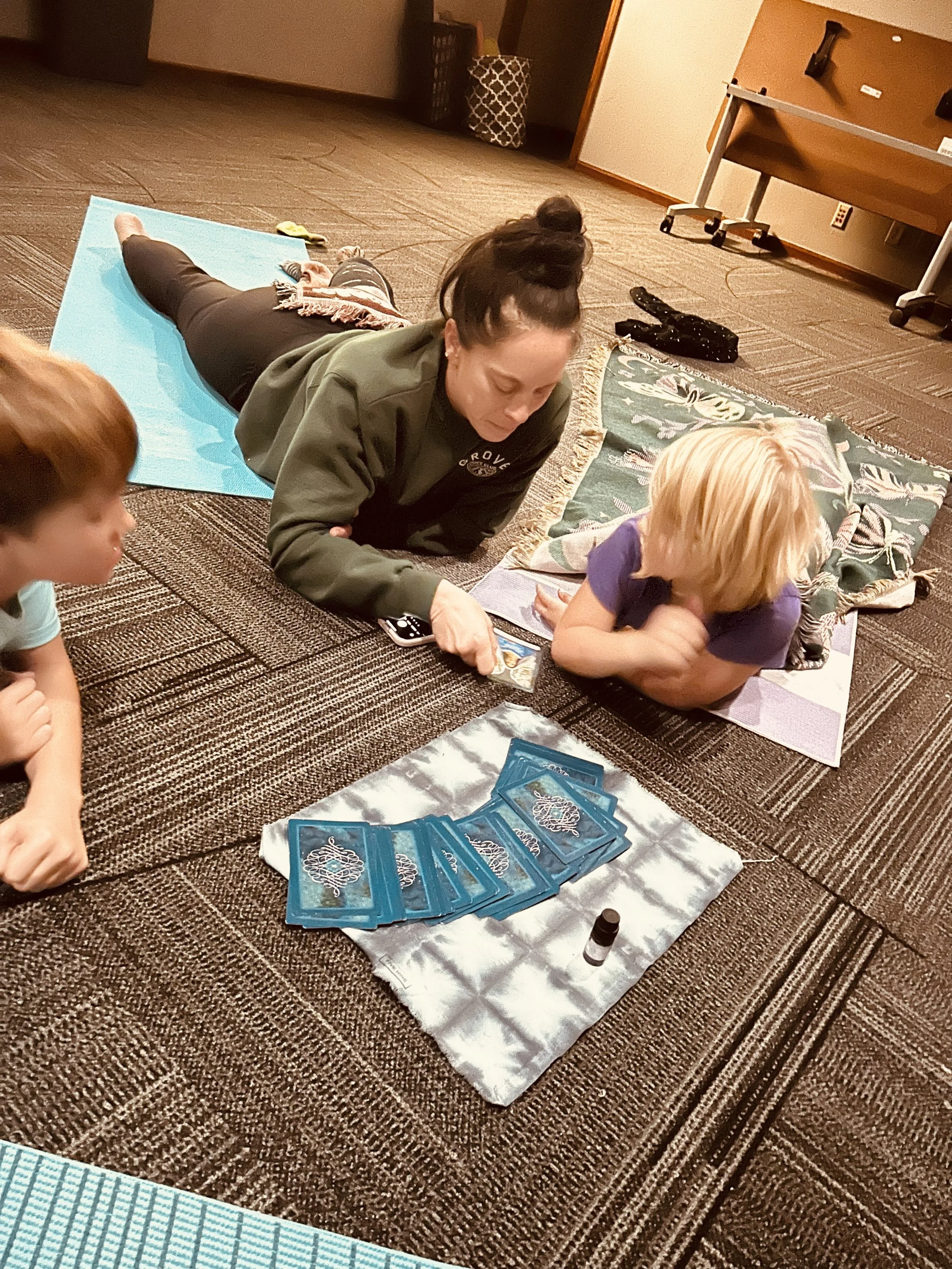A woman and a child lying on the carpeted floor, engaged in a card game with blue tarot cards. There are additional cards, a bottle of essential oil, a gray item, and yoga mats nearby, in a room with a table and some baskets.