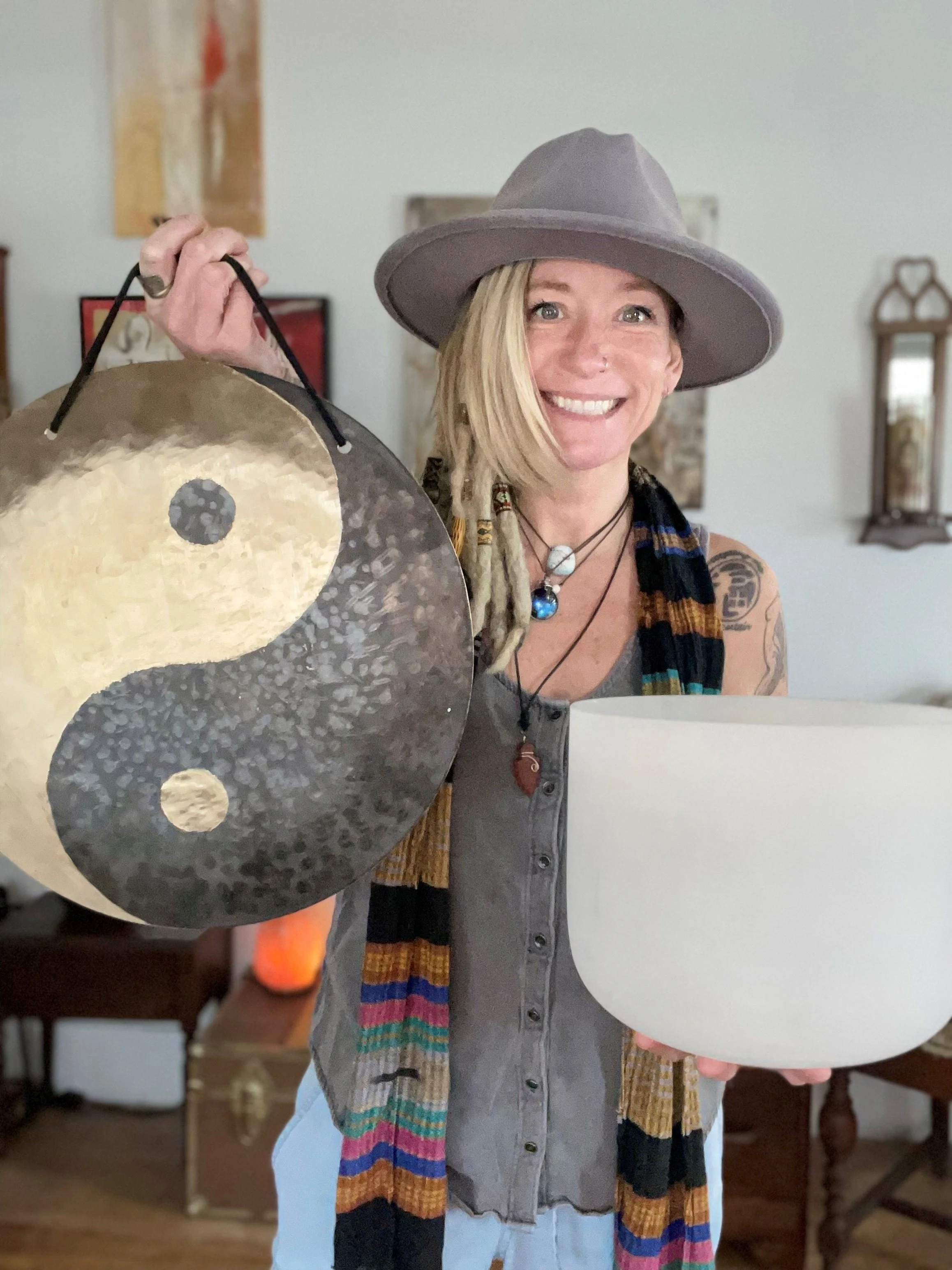 Woman with blonde hair, wearing a gray hat, colorful striped scarf, gray vest, and necklace, smiling while holding a large gong and a white cup, in a home with a mirror and artwork on the wall.