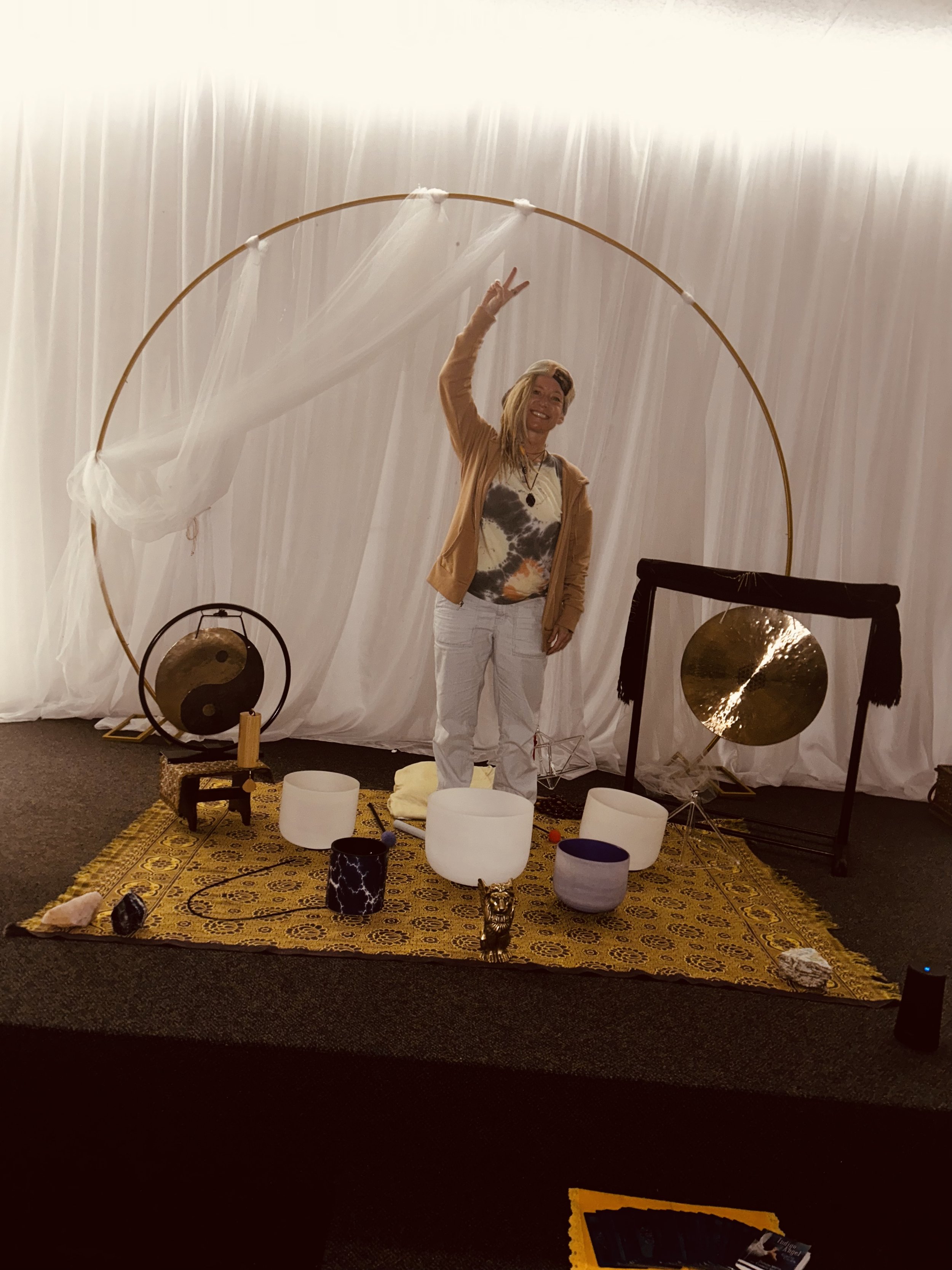 A woman standing inside a decorative setup with a large golden circle, white sheer fabric, and various musical instruments including singing bowls, gongs, and a small drum on a yellow patterned rug.