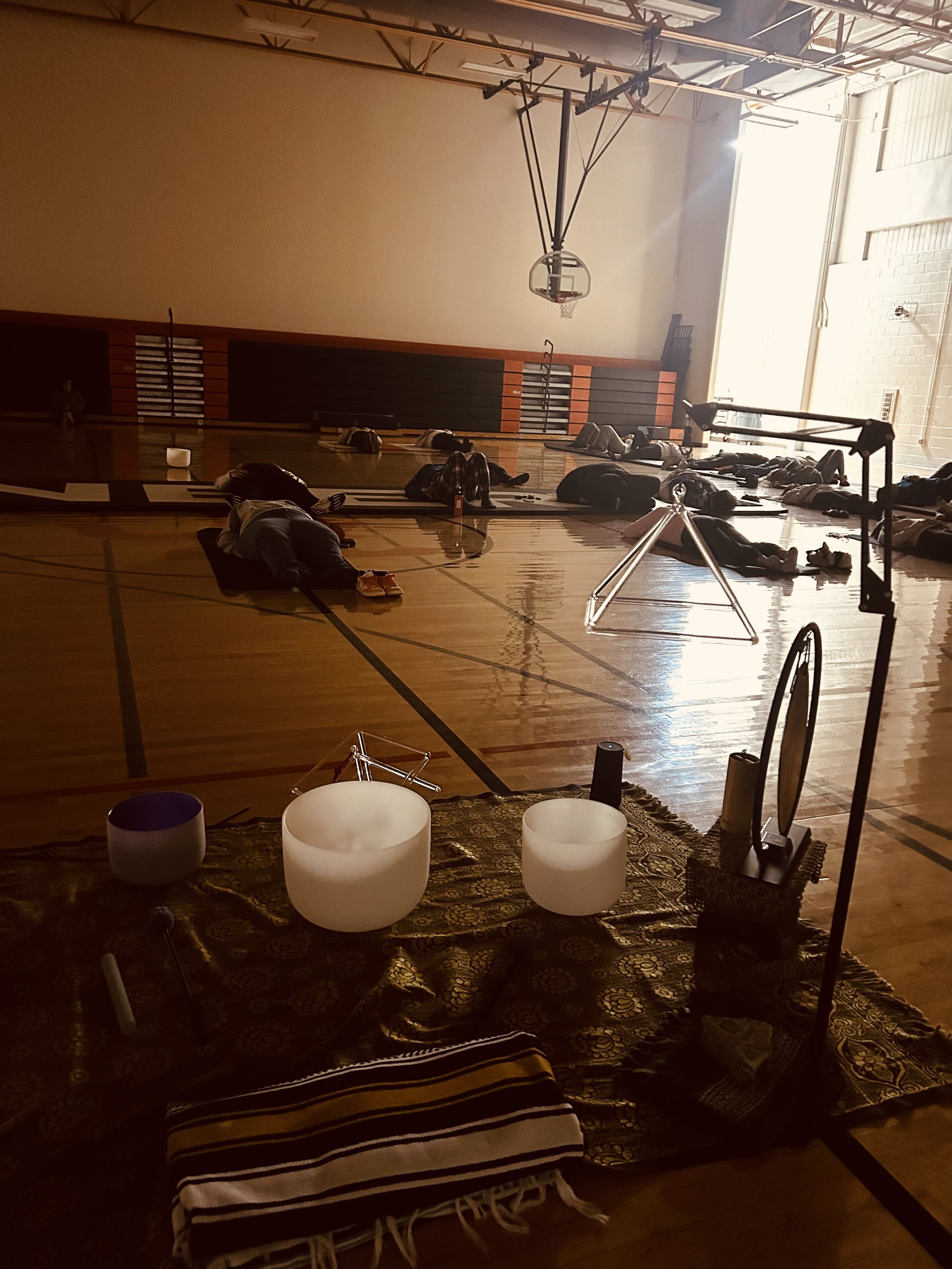A gymnasium with people lying down on yoga mats, some with their eyes closed, participating in a relaxation or meditation session. There are three white singing bowls on a patterned rug in the foreground, and professional photography equipment is set