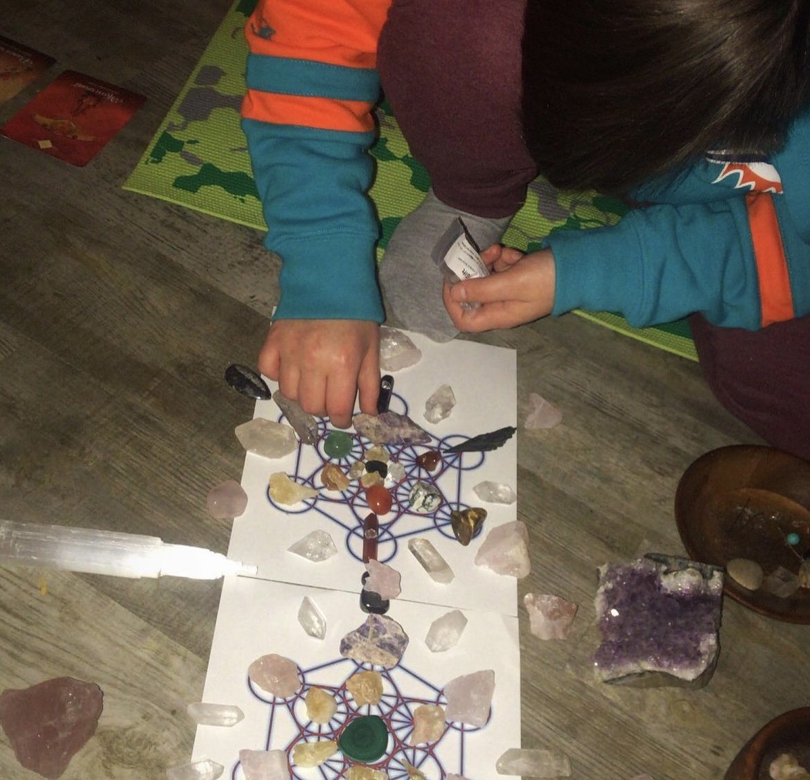 A person is working with crystals and gemstones on two sheets of paper featuring complex geometric patterns, surrounded by various crystals and gemstones on a wooden table.