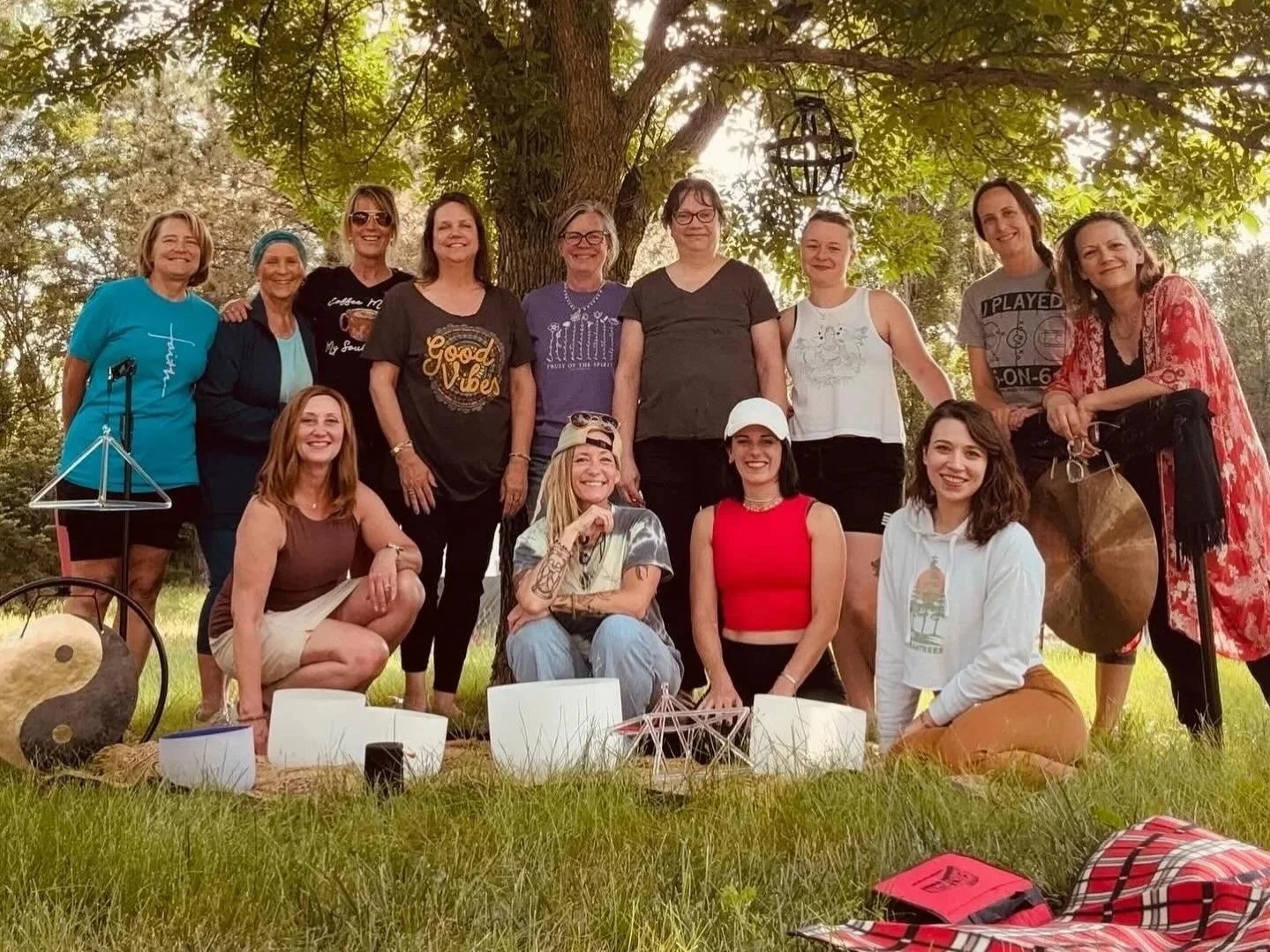Group of people posing outdoors under a large tree, some standing and some kneeling, with musical instruments and decorative items in front, during a sunny day.