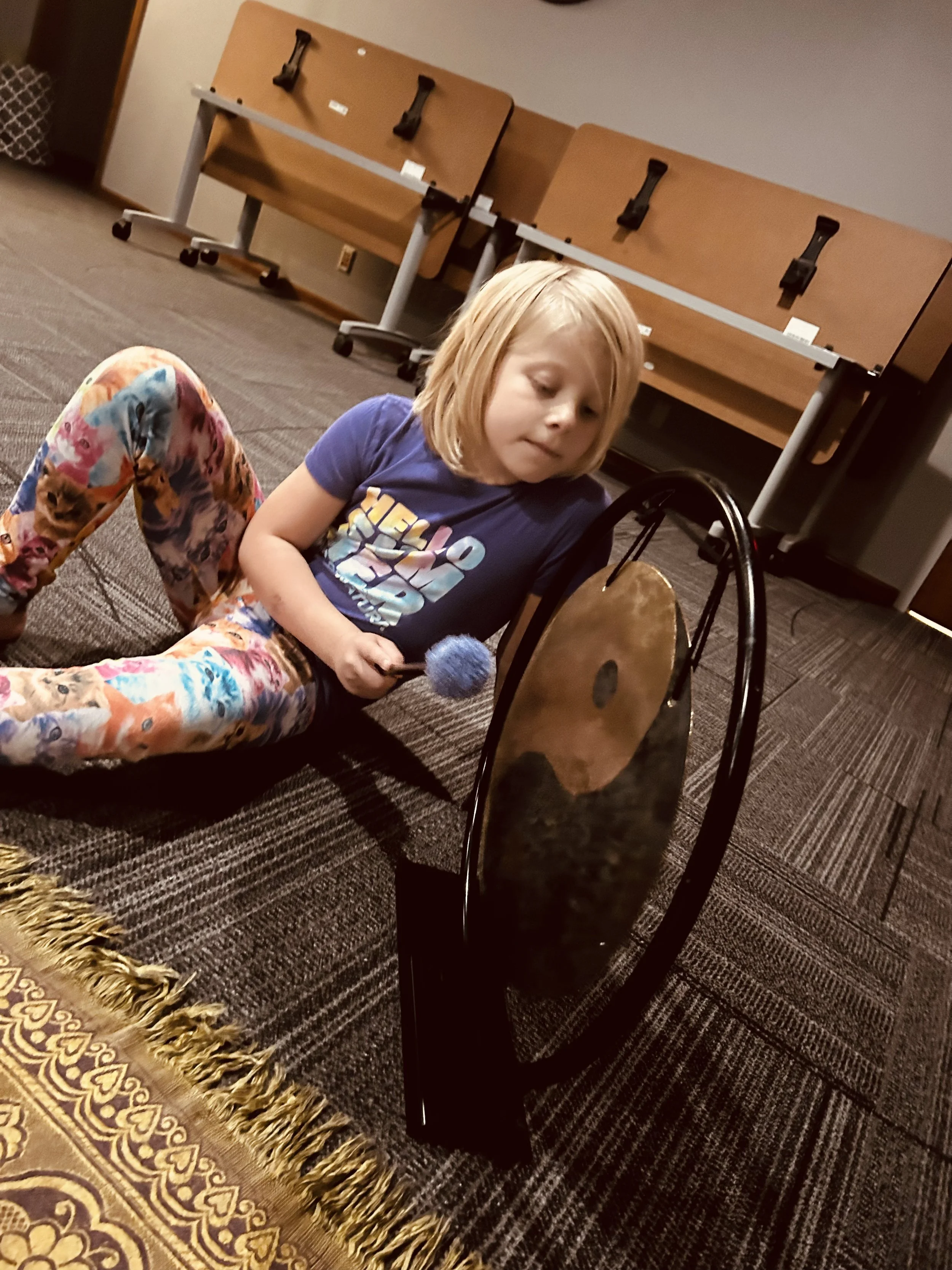 A young girl with blond hair lying on a carpeted floor, holding a blue pom-pom, in front of a brass cymbal, with a brown table flipped upside down in the background.