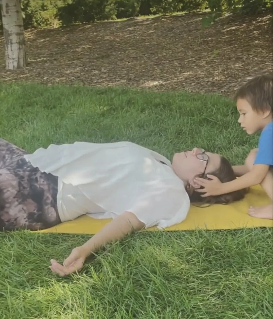 A young girl is lying on a yellow mat outdoors on grass, with a young boy kneeling beside her, holding her head gently.