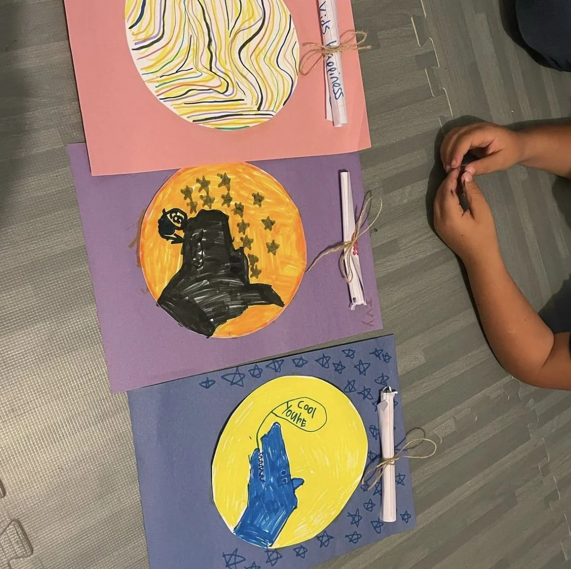 Colorful handmade paper crafts with drawings and messages, displayed on mats, and a child's hands resting nearby.
