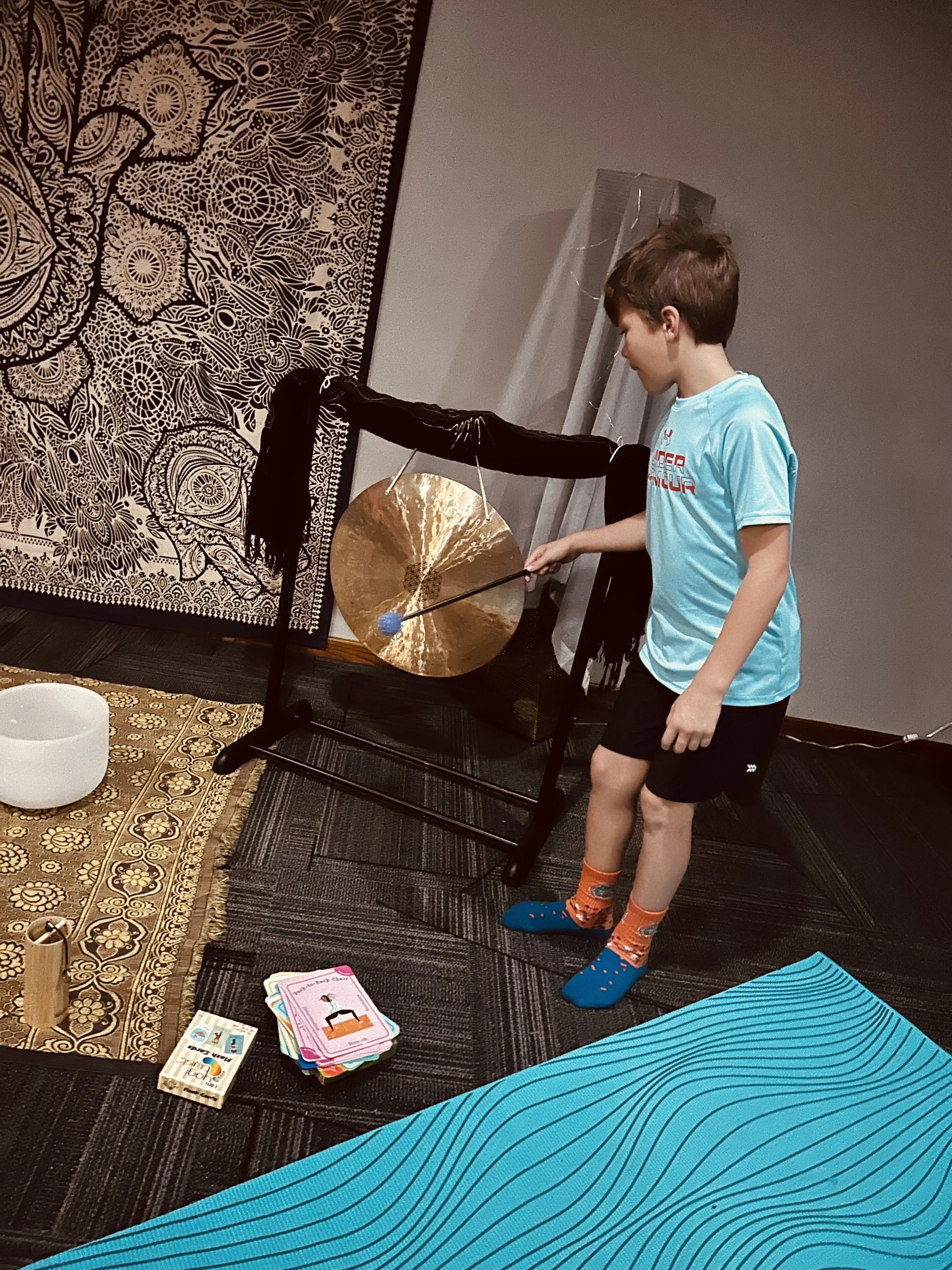 A young boy in a blue t-shirt and colorful socks stands on a yoga mat, holding a mallet and hitting a gong in a room with a detailed tapestry on the wall, books, and yoga equipment.