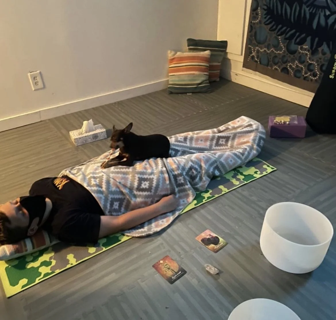 A person lying down on a yoga mat with a blanket over them, a small black cat lying on its back on top of the blanket, in a room with a gray floor, pillows, a box of tissues, and a white singing bowl.