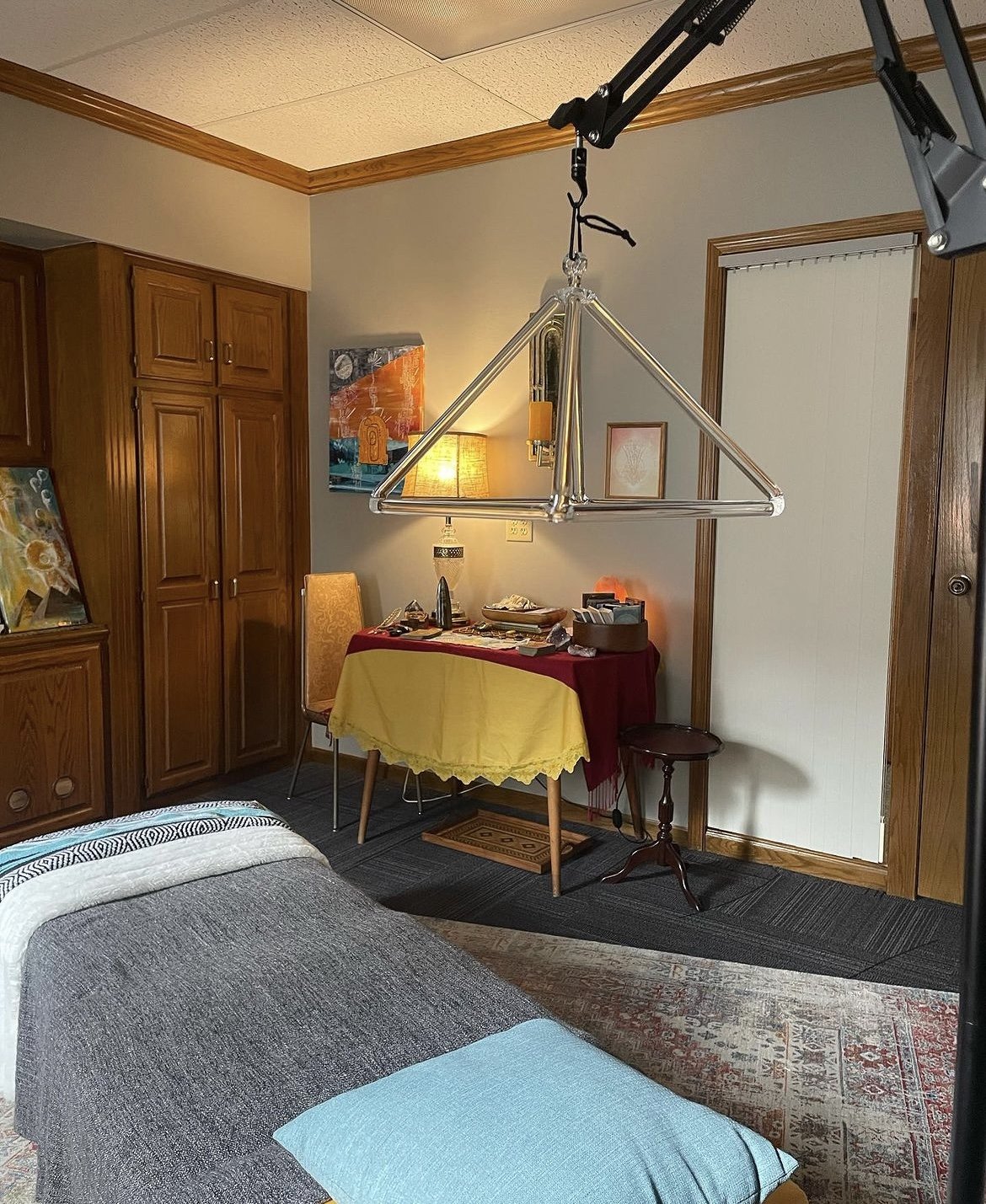 A hotel room with a bed, a corner table with a yellow tablecloth, a wooden cabinet, and a ceiling-mounted trianglular light fixture.