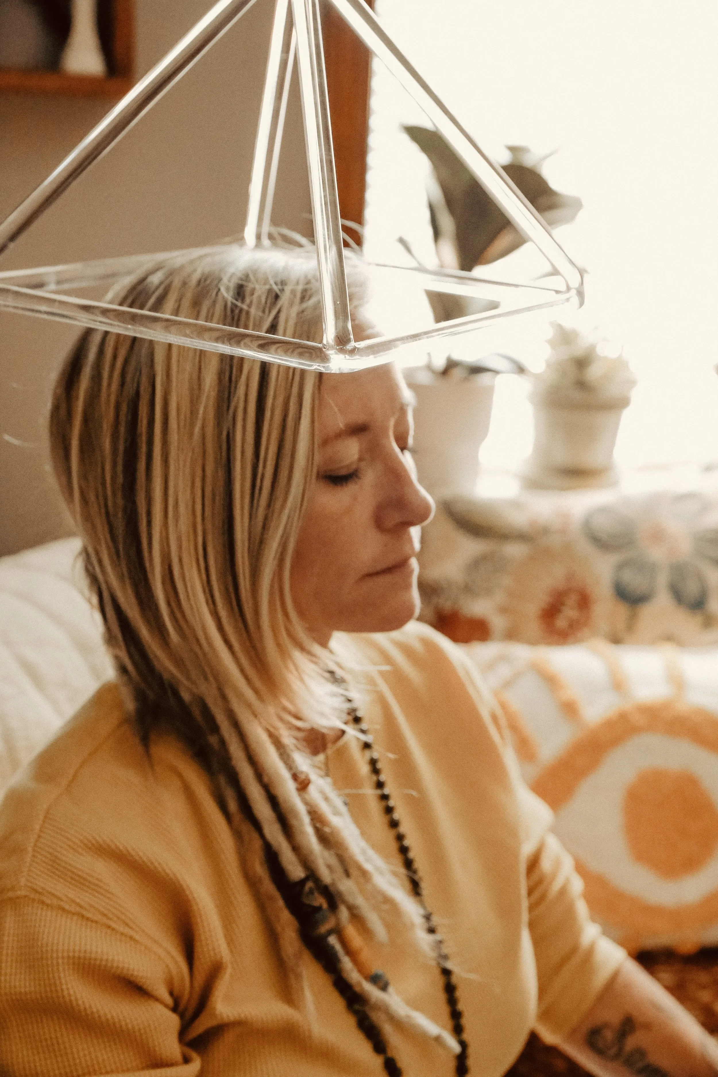 A woman with blonde hair and tattoos on her arm is sitting with her eyes closed. There is a geometric glass sculpture balanced on her head, and in the background, there are potted plants and a floral patterned object.