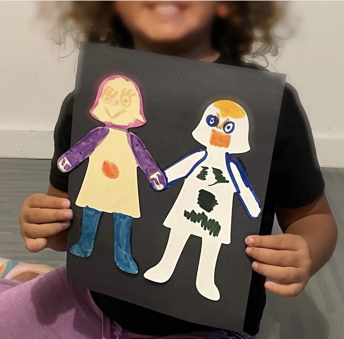 Child holding a black poster with colorful paper cutouts of a girl and a bird. The girl has pink hair, purple sleeves, and blue boots. The bird has a white body, orange beak, and blue eyes, with green patches on its chest.