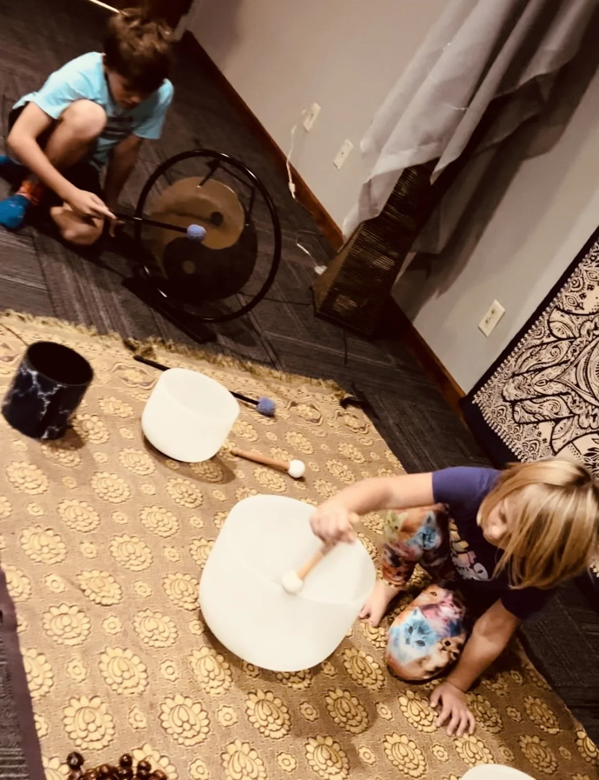 Two children playing a sound therapy game with mallets and singing bowls on a patterned carpet, in a room with a curtain, a wall with multiple electrical outlets, and a stand with hanging beads.