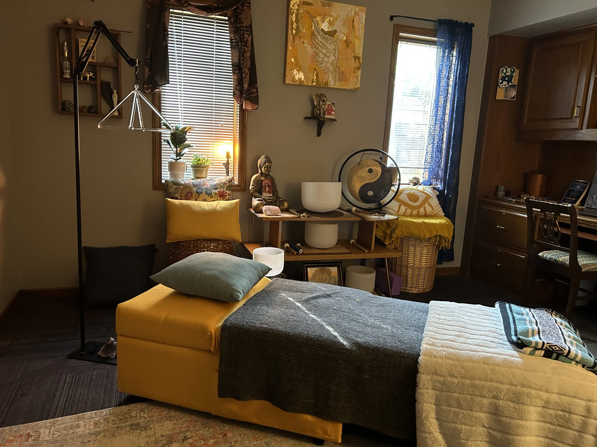 Sound Healing room for Sound Baths. Living room with a yellow sofa, assorted pillows, a window with blinds and floral curtains, a Buddha statue, a singing bowl, and spiritual decorations on a small table.