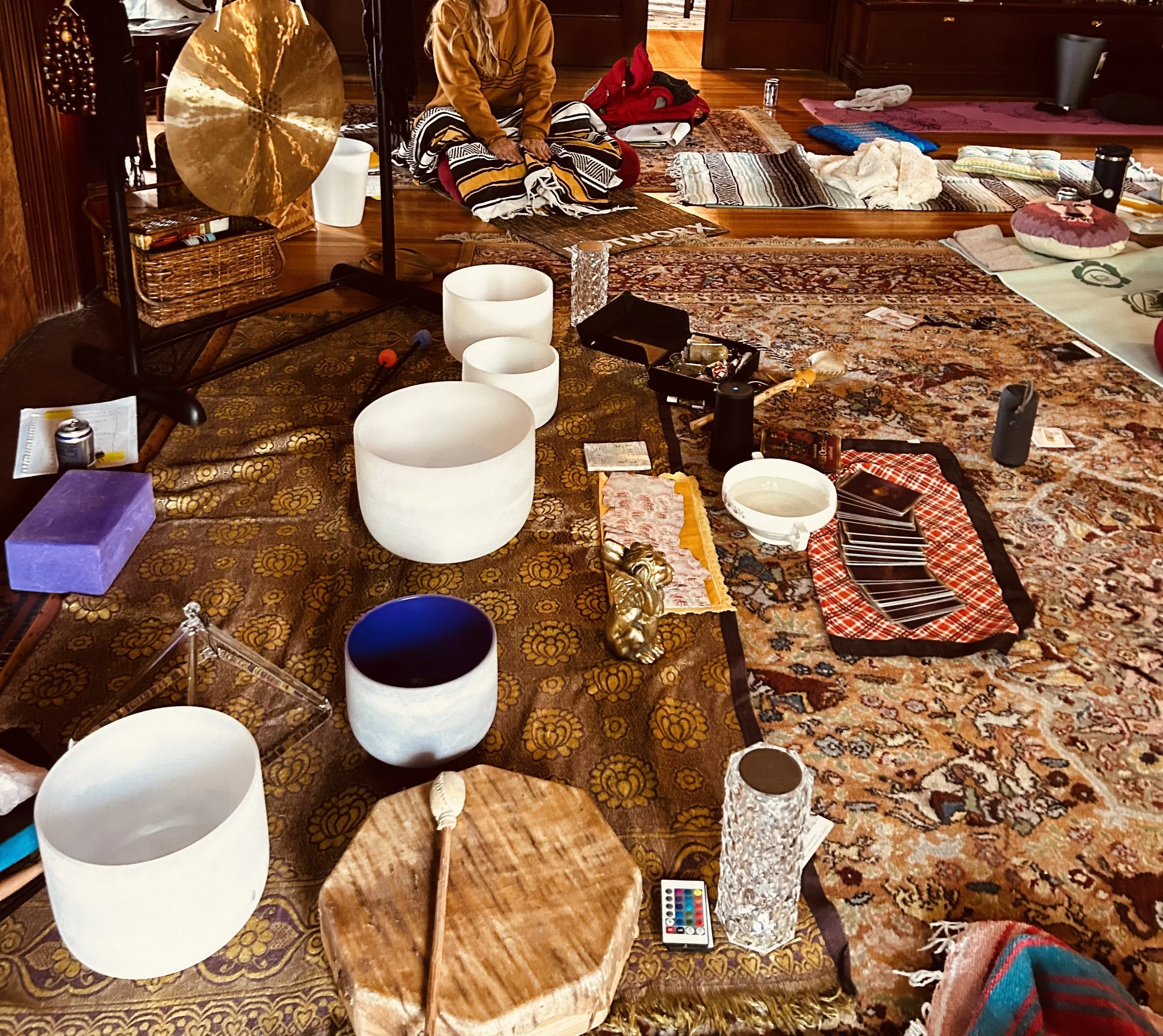 A room with a patterned rug and a variety of objects scattered on the floor, including white singing bowls, a purple box, a small mirror, wrapped items, a small basket, and papers. There are mats and cloths laid out, and a person sitting cross-legged