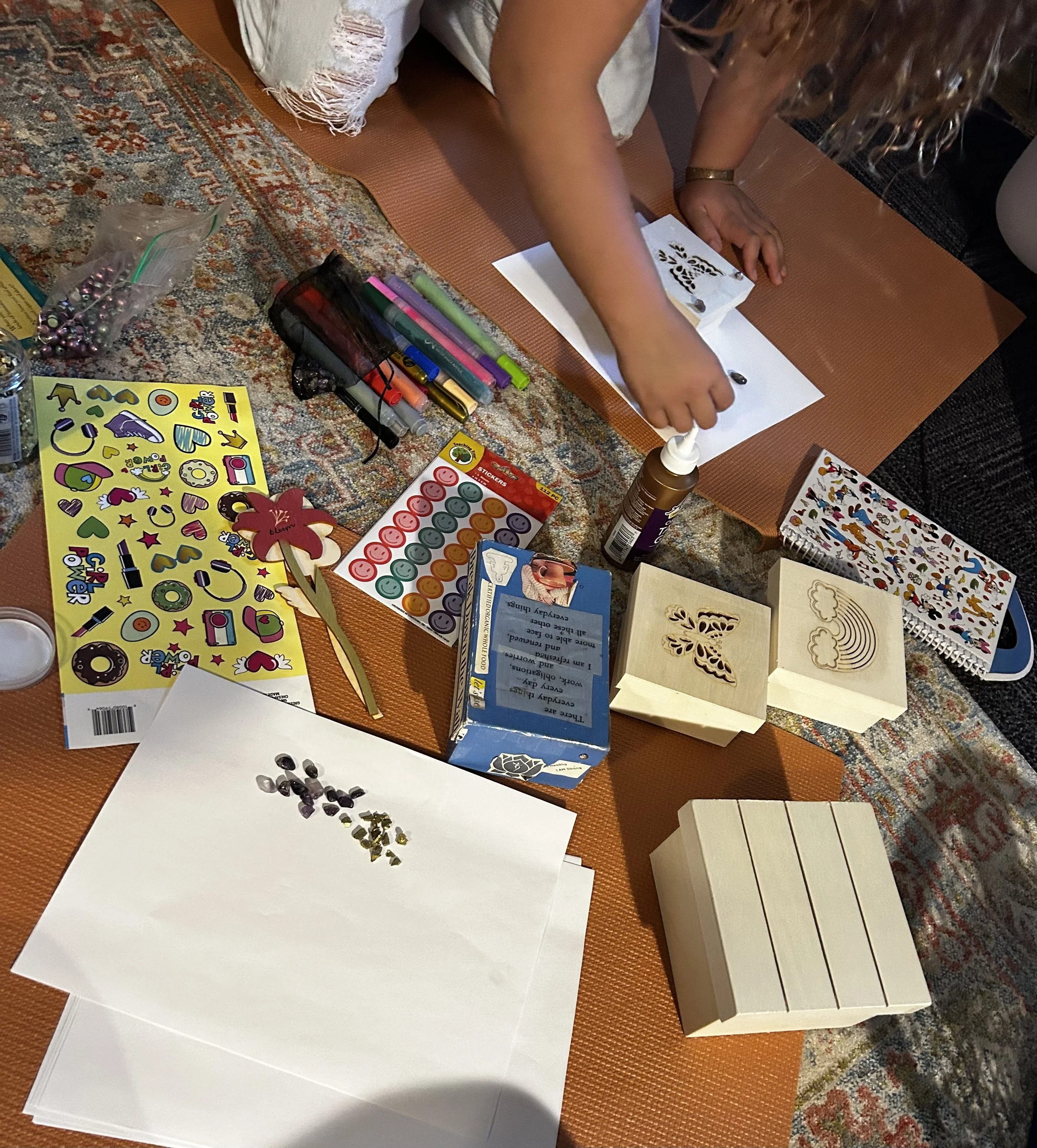 A person working on a scrapbooking or craft project on a table, surrounded by various art supplies like markers, stickers, decorative paper, and wooden blocks. The person is using a stencil or stamps on a piece of paper.