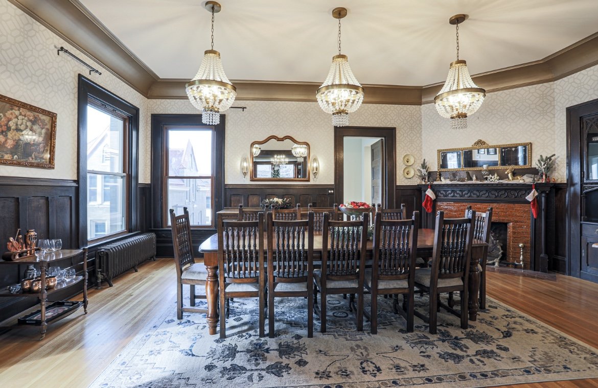 Elegant dining room with dark wood paneling, a large wooden table surrounded by ten chairs, an ornate fireplace with stockings and holiday decorations, a large window, and three chandeliers hanging from a ceiling. There is a framed painting on the wall, a mirror, and a side table with glassware and copper decorations.