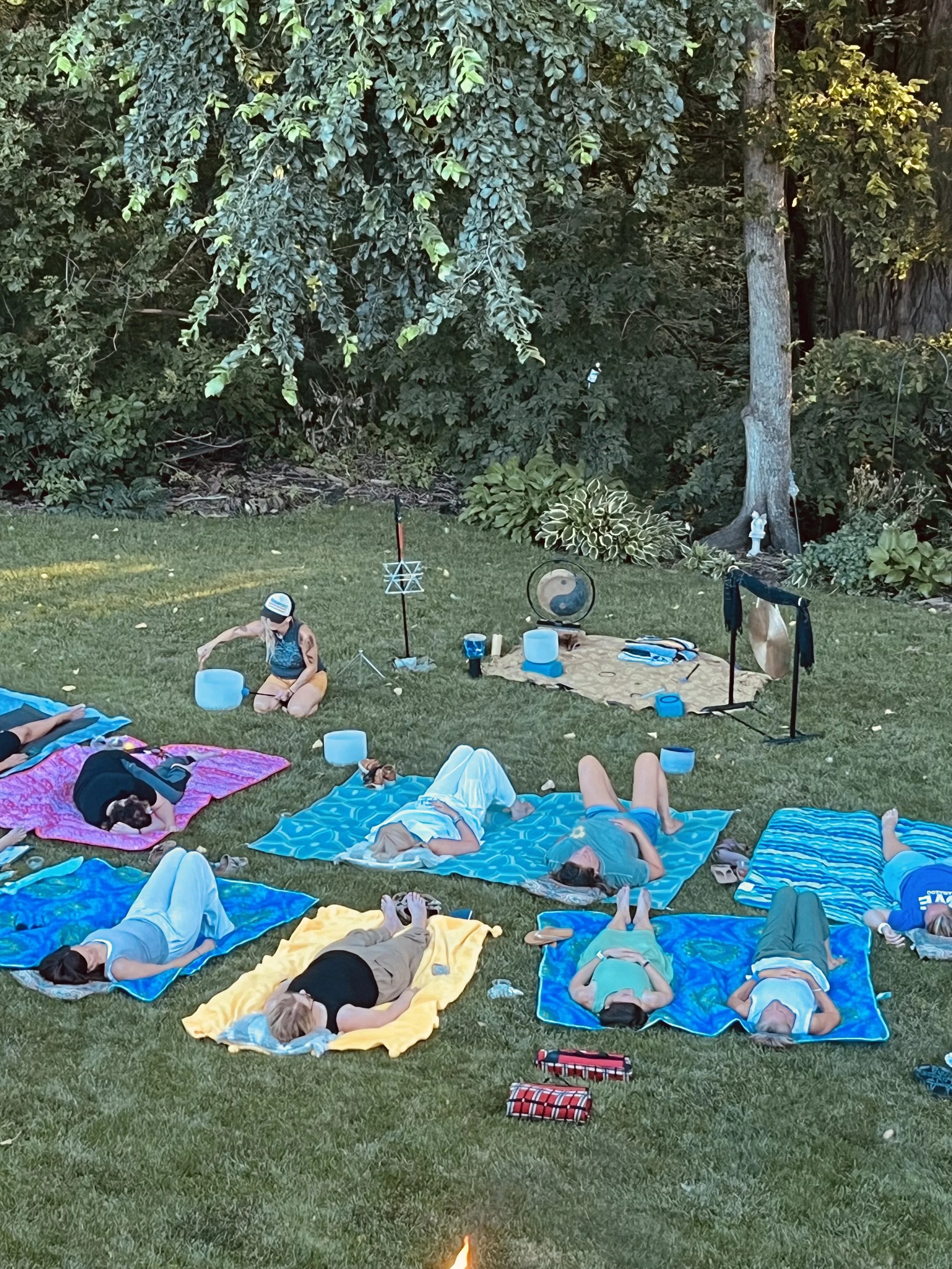 People lying and meditating on colorful towels in a backyard with grass, trees, and garden statues.