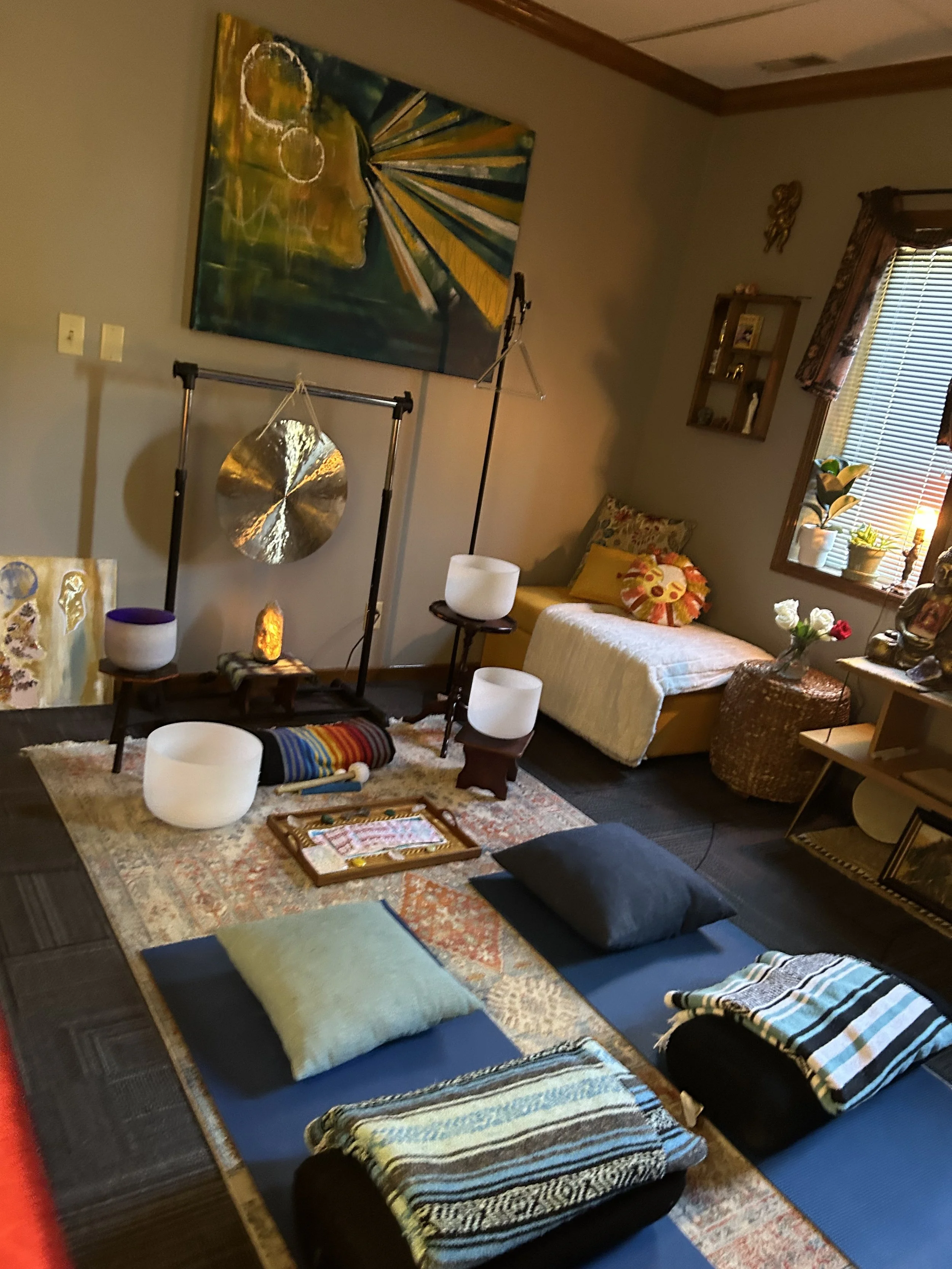 This image shows a serene indoor setting with various meditation cushions and mats laid out on the floor, suggesting a space dedicated to relaxation or meditation. There are decorative elements including a large ceramic bowl, a plate, and a book on a