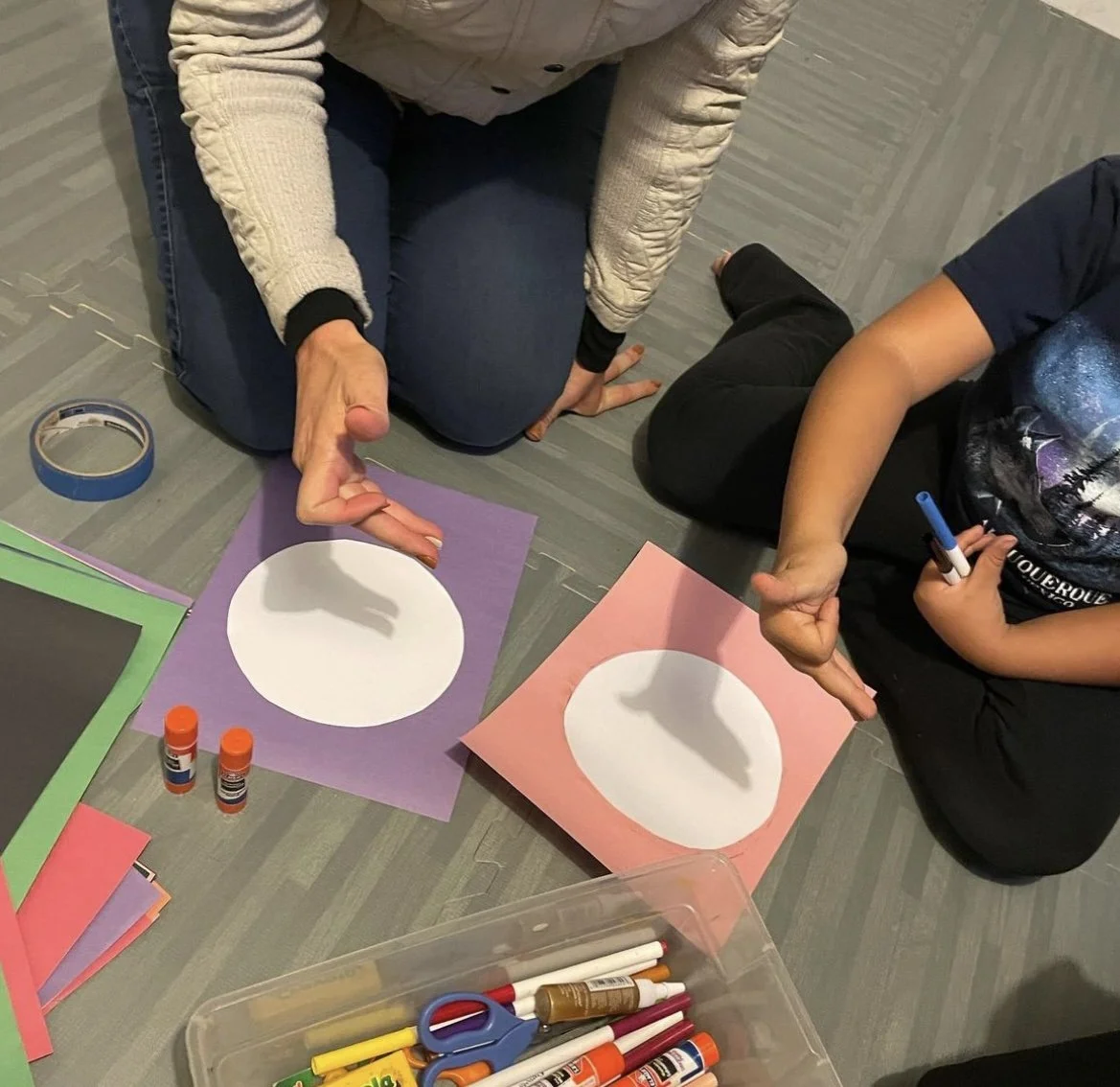 Two children and an adult sitting on the floor, working on an arts and crafts project with colored paper, glue sticks, scissors, markers, and tape. The adult is demonstrating a craft with their hand near the paper, which has a large white circle on p