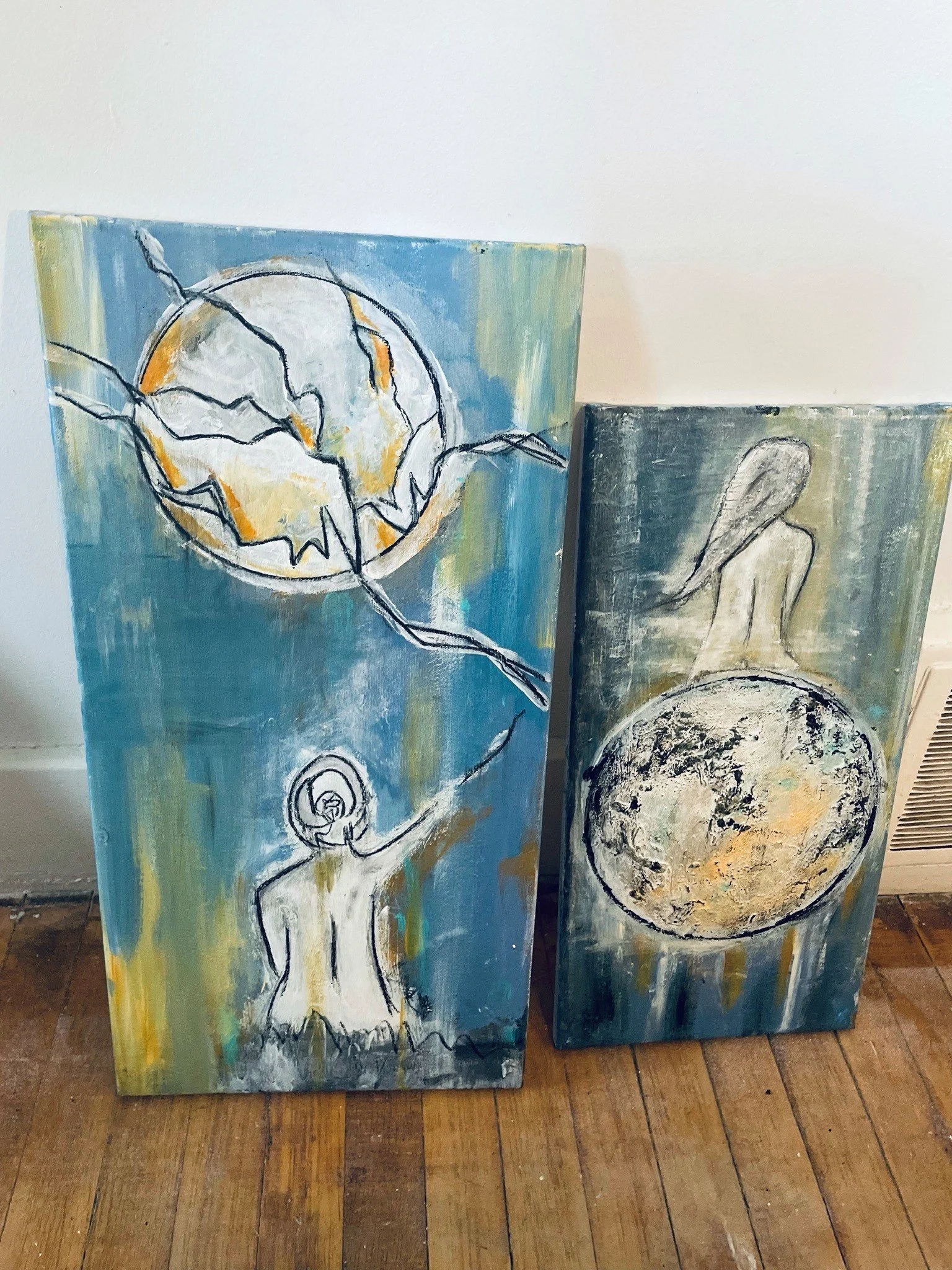 Two paintings on wooden panels leaning against a white wall; one features an abstract woman with a large spherical head and long limbs, the other depicts a coin on a dark background.