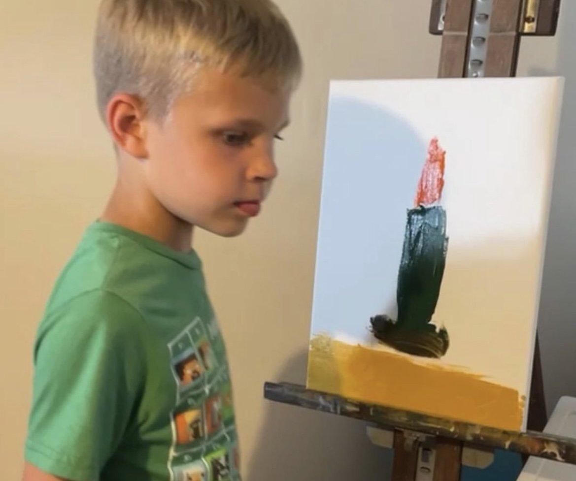 A young boy with blonde hair wearing a green t-shirt is standing in front of a canvas, focusing on a painting with broad, bold strokes of red, black, yellow, and green.