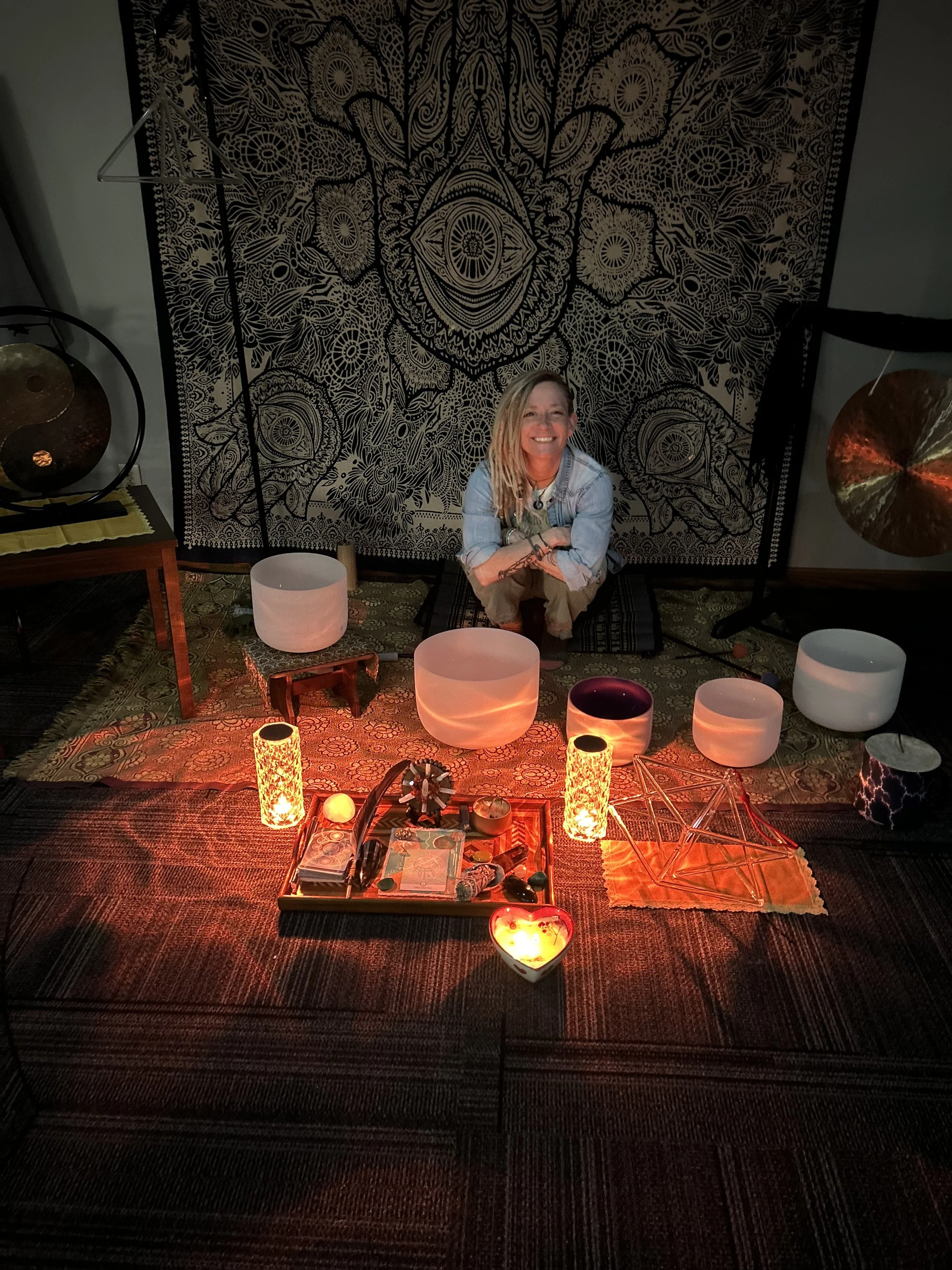 A woman with blonde hair and a smile, sitting on a cushion in a room decorated with spiritual and mystical objects, including lit candles, a star-shaped ornament, a heart-shaped candle holder, a hanging tapestry with intricate patterns, and sound bowls, with ambient lighting creating a cozy and spiritual atmosphere.