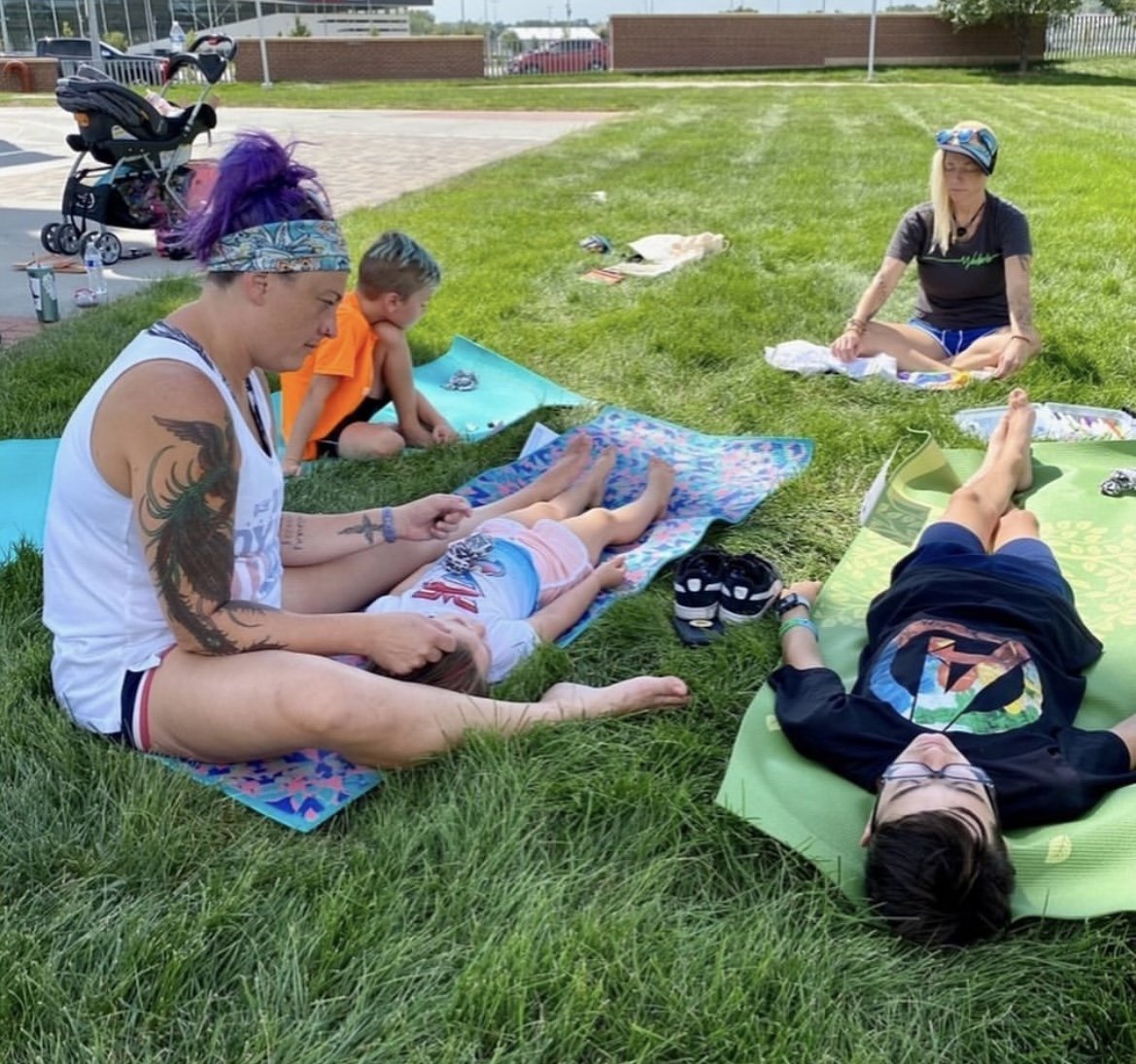 Group of people relaxing on yoga mats on the grass in a park, with children and adults sitting and lying down. A stroller, water bottles, and personal items are visible nearby.