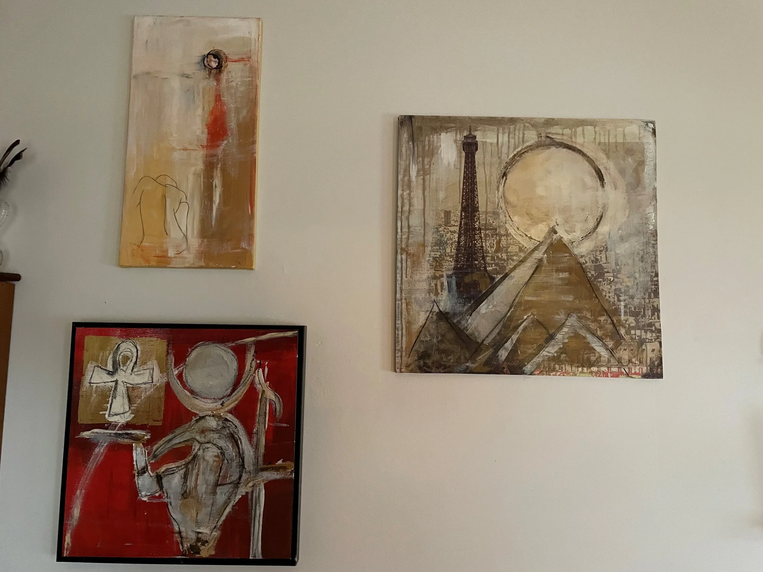 Three abstract paintings on a wall, with colors including red, brown, beige, and white. The central painting features a landmark resembling the Eiffel Tower in Paris, with a circular element and a pyramid shape at the bottom.