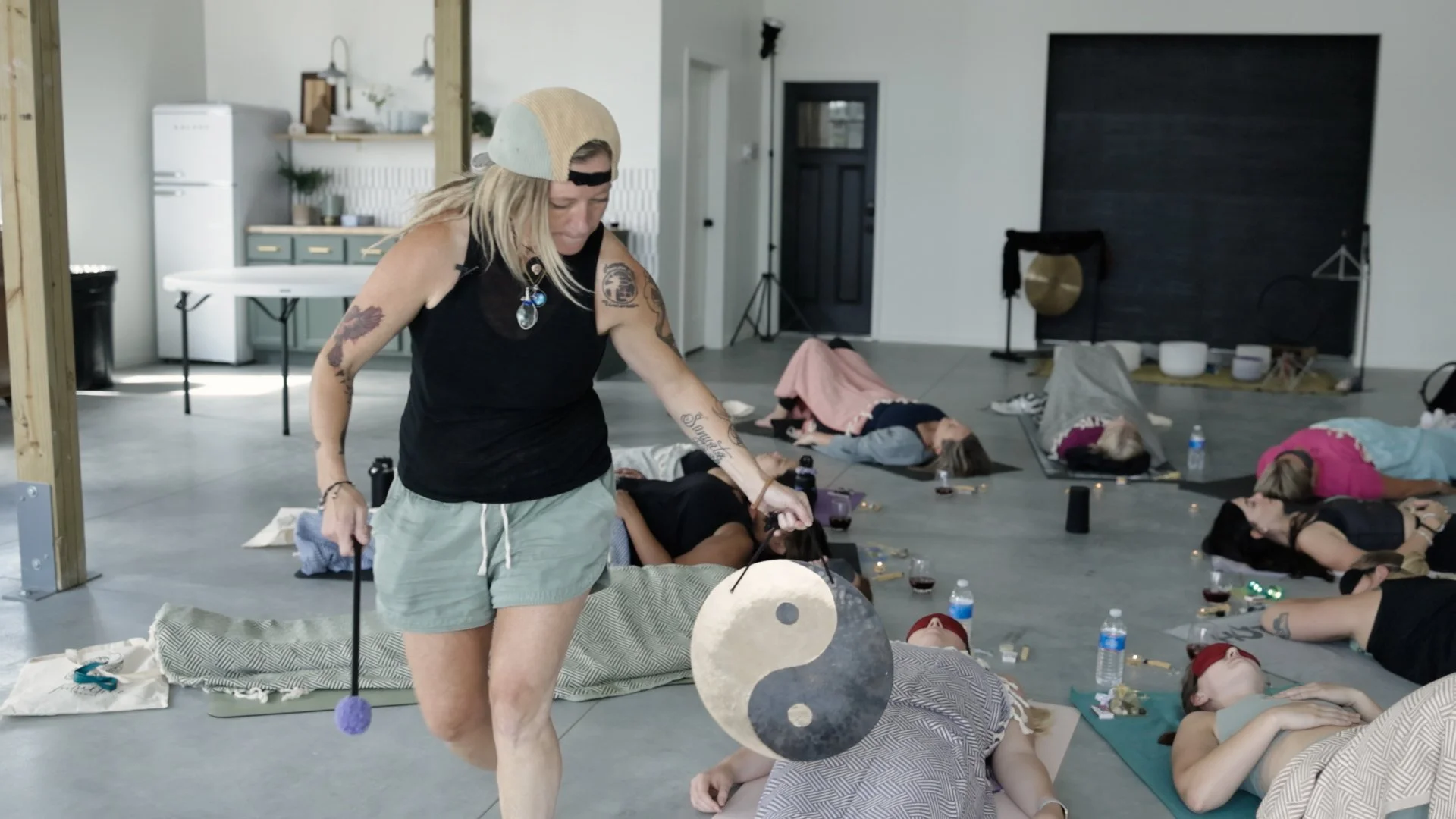 A woman standing in a yoga or meditation class holding a gong in a large room with several people lying on mats, some with blankets, and water bottles nearby. The room has a minimalistic decor with a blackboard and musical instruments in the backgrou