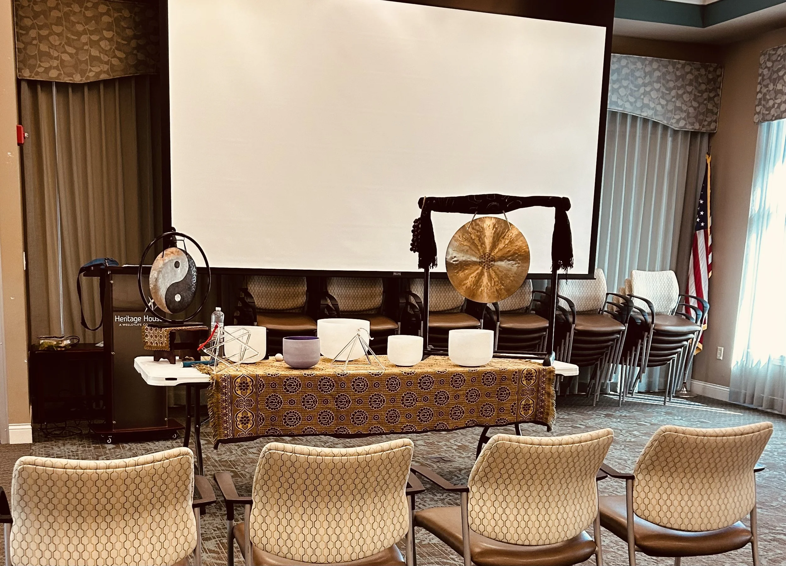 A conference room with chairs arranged in rows and a large projection screen at the front. The room has patterned curtains, a table with Tibetan bowls, a gong, and musical instruments, as well as an American flag near the window.