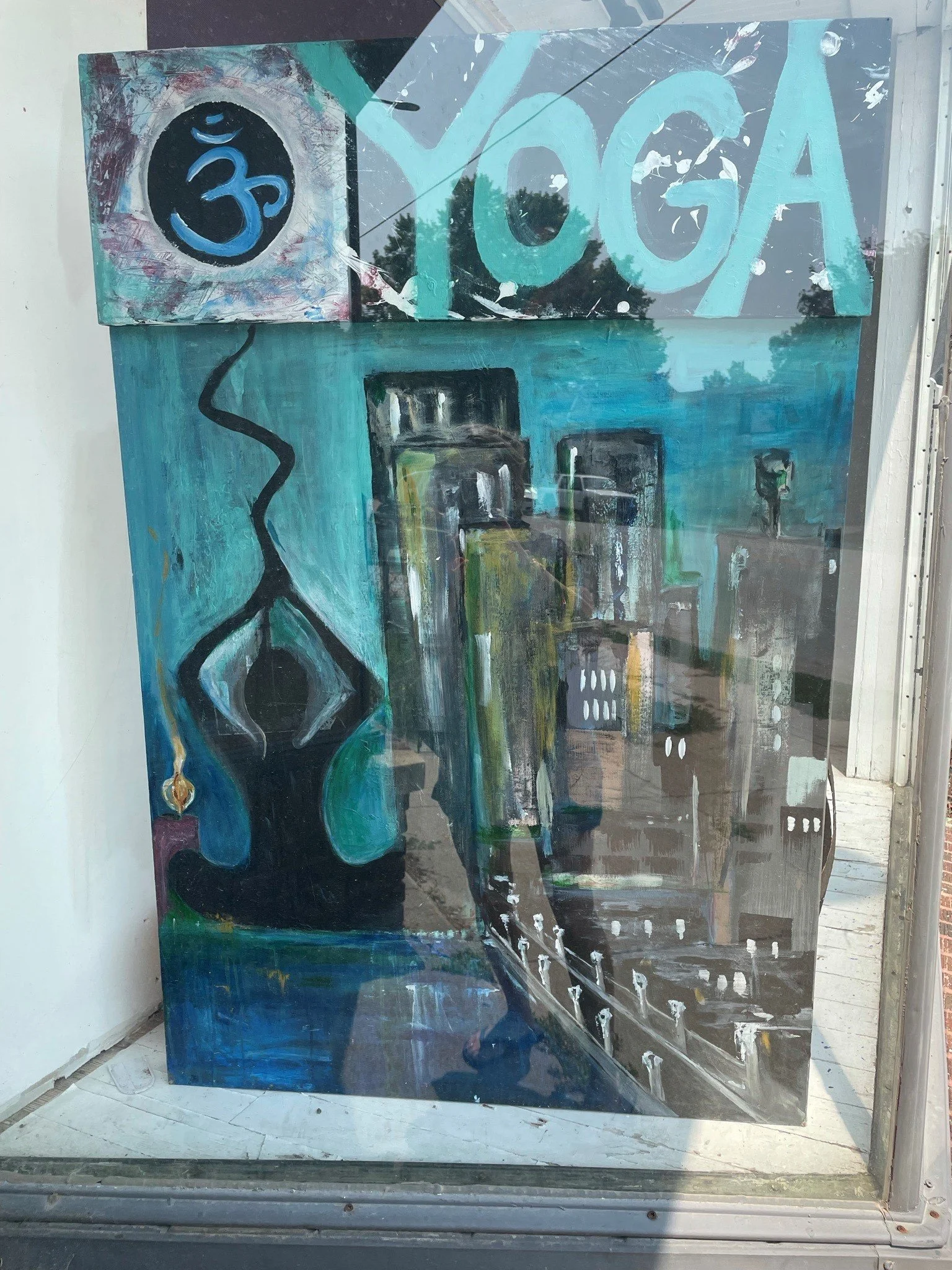 A colorful abstract painting with a prominent turquoise OK sign and a black circle with a blue Om symbol inside it, displayed behind glass with reflections of trees and sky.