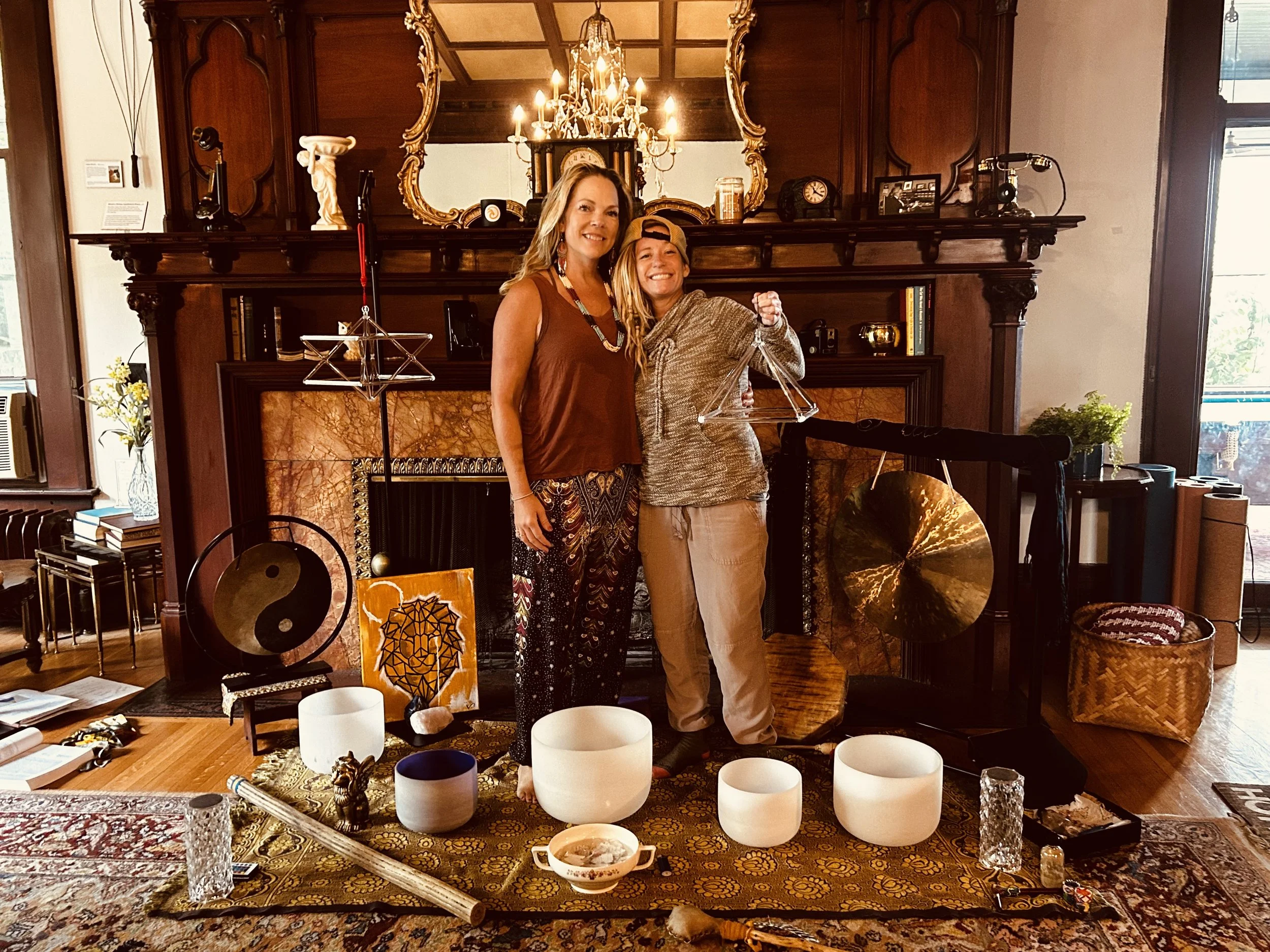 Group Sound Bath Retreat. Two women, one older and one younger, standing in a living room with a large ornate mirror, chandelier, and fireplace. They are smiling, and there are various decorations and musical instruments around them.