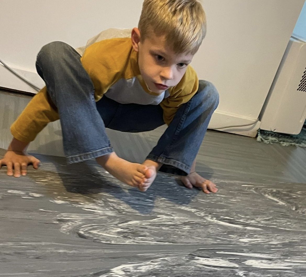 A young boy squats on the floor, playing with a large sheet of plastic that has some white streaks on it.