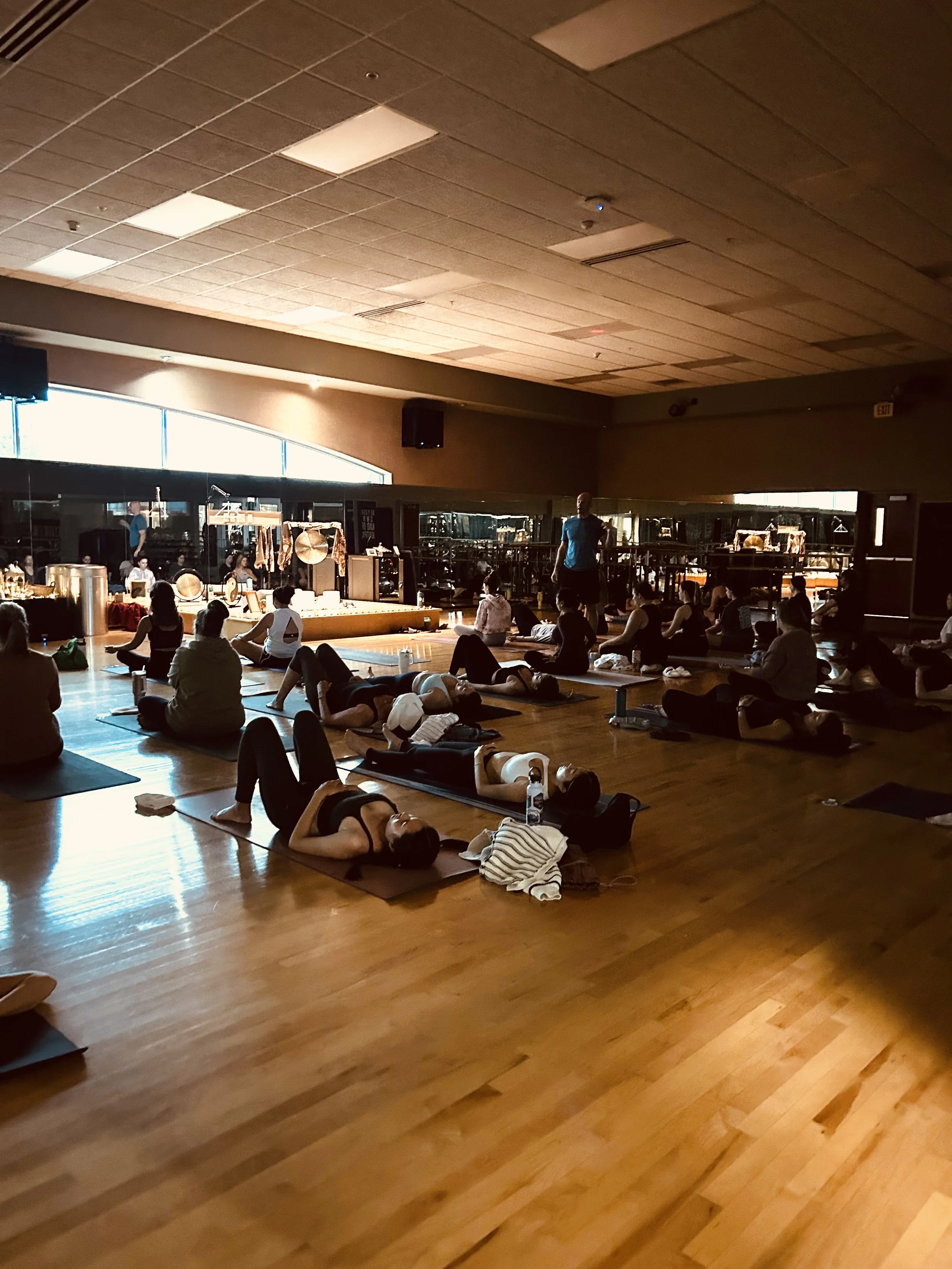 Sound Bath in Omaha, NE. People participating in a yoga or meditation class inside a spacious room with wooden floors and a high ceiling, some sitting or lying on yoga mats