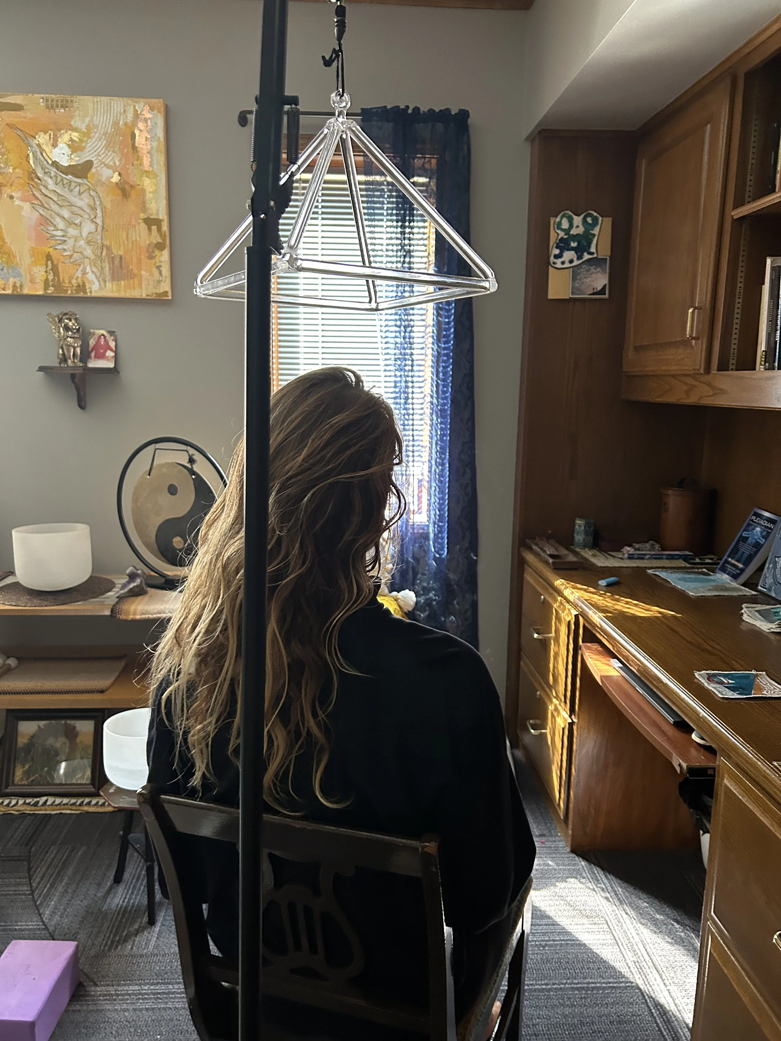 A woman with long, wavy hair sitting at a desk in a room with wooden furniture and decorative items. The room has a window with blinds and blue curtains. There is a geometric lamp hanging overhead and various objects on the desk and shelves.