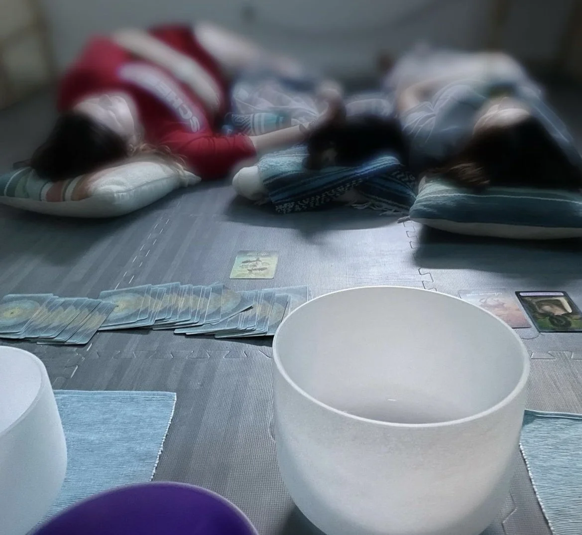 Three children lying on pillows on the floor, resting or sleeping. Playing cards and a game board are laid out on the floor in front. The scene appears to be indoors with a casual, relaxed atmosphere.
