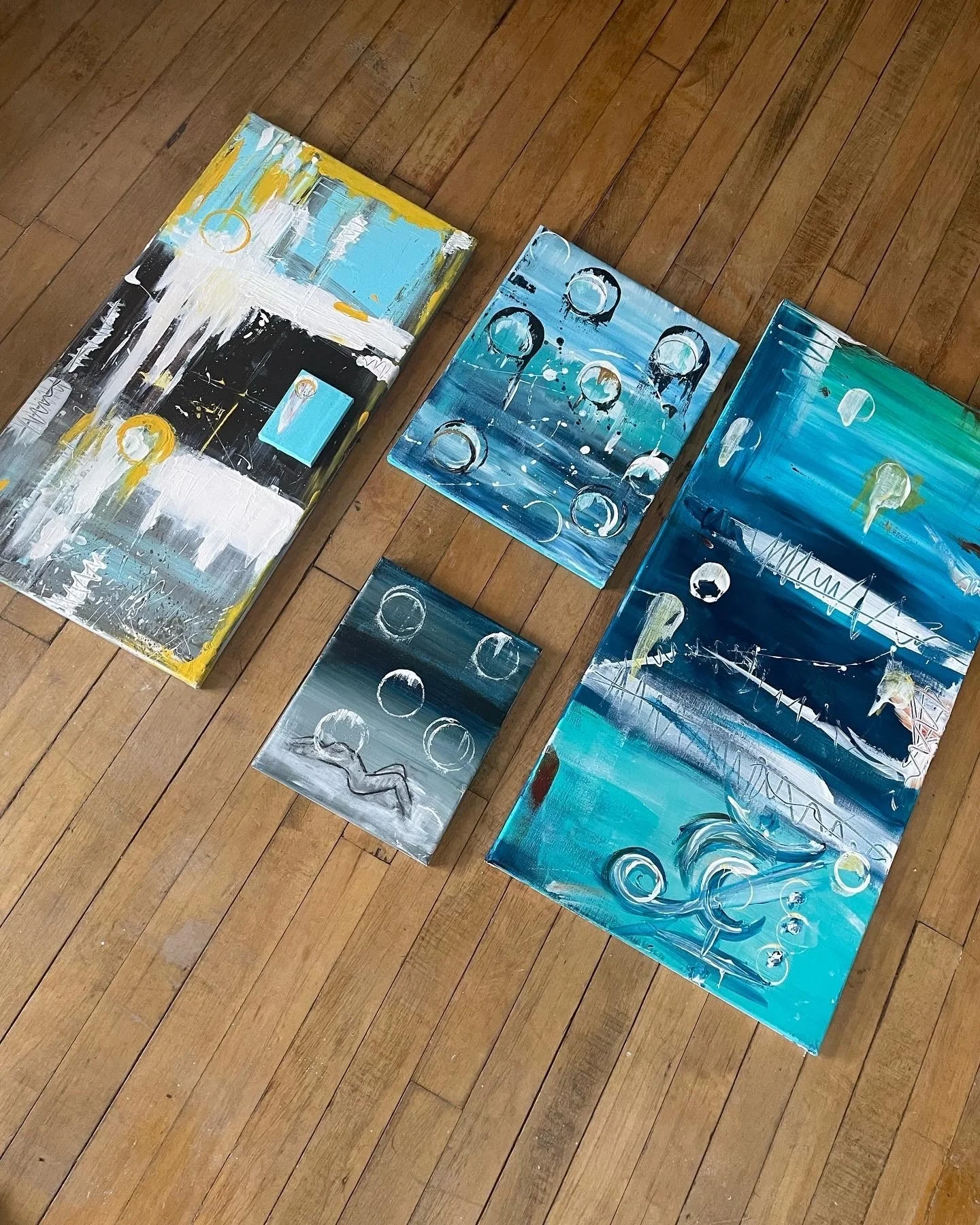 Four abstract paintings with blue, black, white, and yellow colors lying on a wooden floor. The paintings feature circular, linear, and splash patterns.