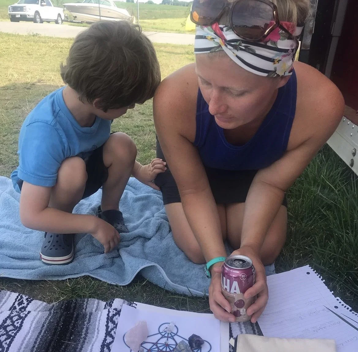 A woman and a young boy sitting on a blue towel outdoors on grass, looking at objects and papers on the ground, with a car and boat in the background.