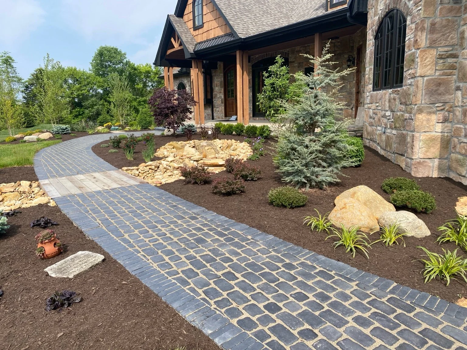 A front yard landscape featuring a cobblestone walkway leading to a house with a brick and stone facade. The garden includes shrubs, small trees, rocks, and a variety of plants with mulch coverage. The house has a covered porch and is surrounded by green grass and trees.
