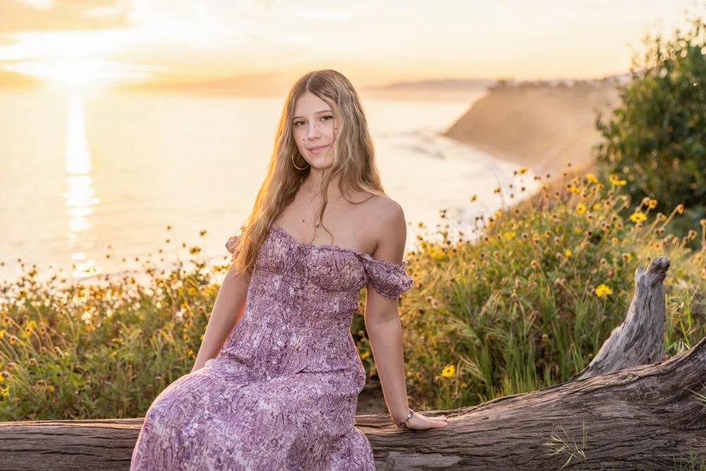 A young woman sitting on a fallen log near the coast during sunset, wearing a purple off-shoulder dress, with yellow flowers and green bushes in the background.