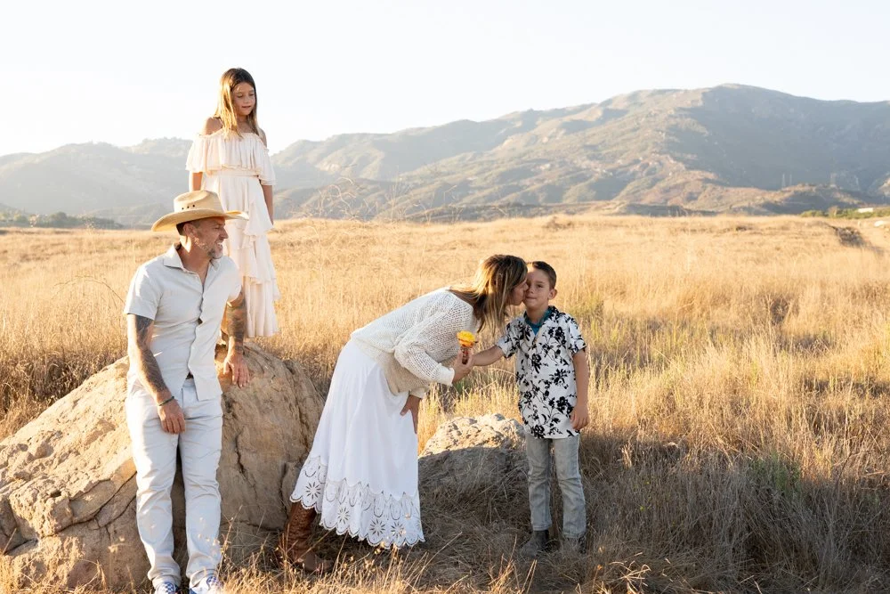 A woman is kissing a young boy on the cheek in a field with mountains in the background, while a girl stands on a rock nearby and a man watches smiling.