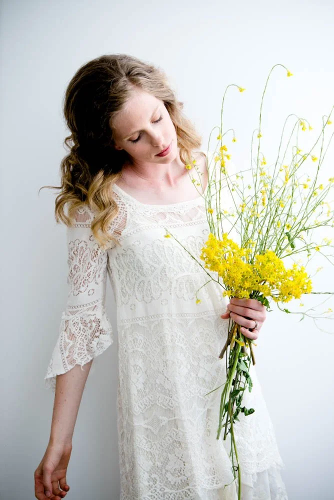A woman with light brown curly hair is holding a large bouquet of yellow flowers and looking down at it, wearing a white lace dress with three-quarter length sleeves against a plain white background.