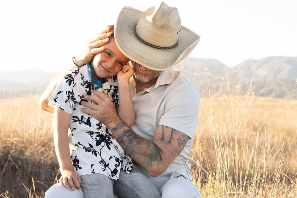 A man and a young girl sharing a moment in a grassy field, with the man wearing a wide-brimmed hat and the girl resting her head on his shoulder.