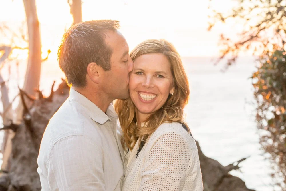 A man kissing a woman on the cheek outdoors at sunset, smiling with the ocean and trees in the background.