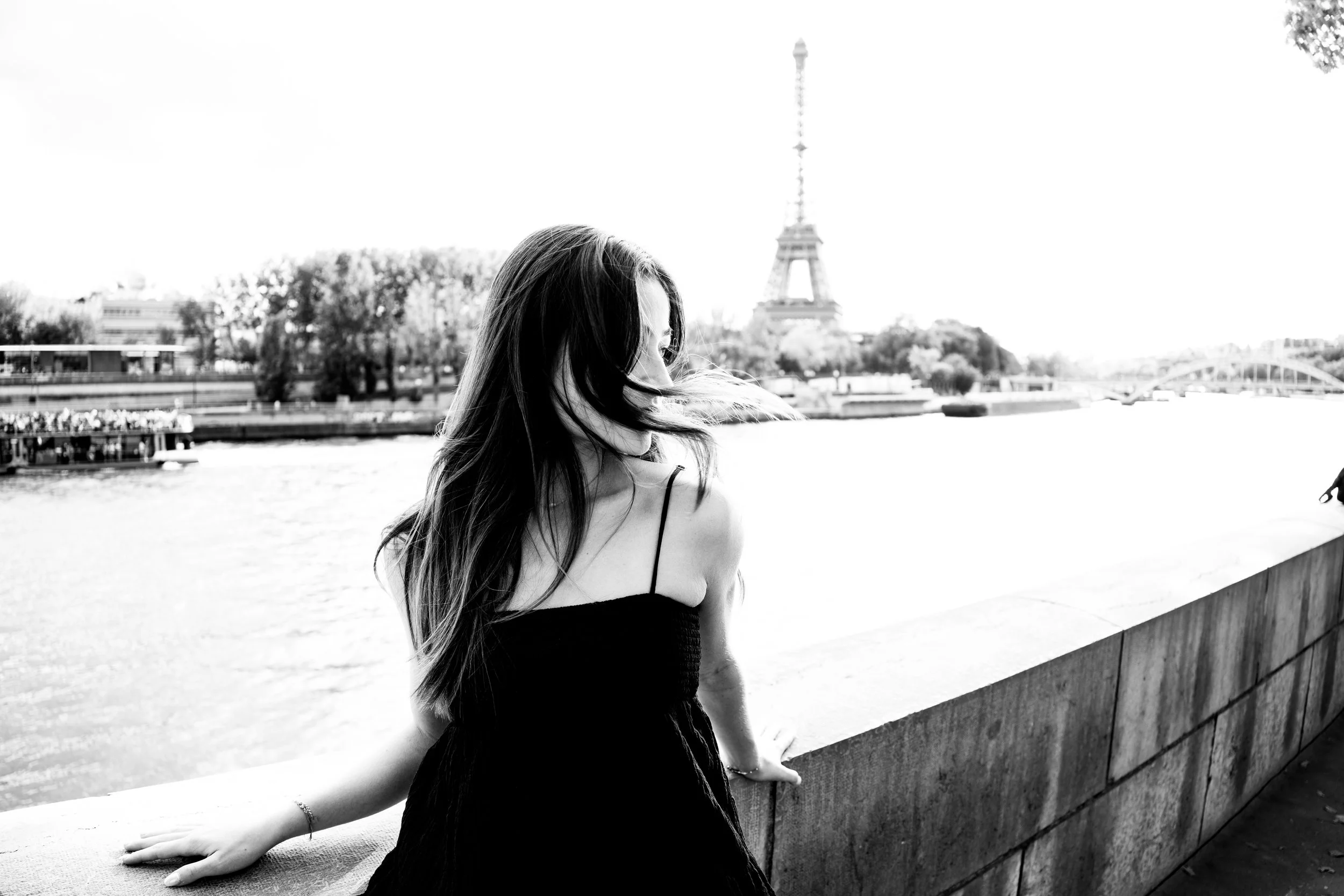 A woman with long hair in a black dress leaning on a stone ledge by the River Seine in Paris with the Eiffel Tower in the background.