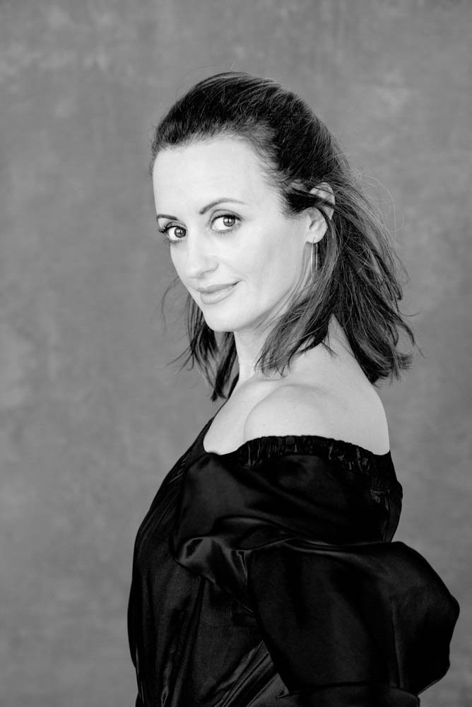 Black and white portrait of a woman with shoulder-length hair, wearing an off-the-shoulder top, looking over her shoulder at the camera.