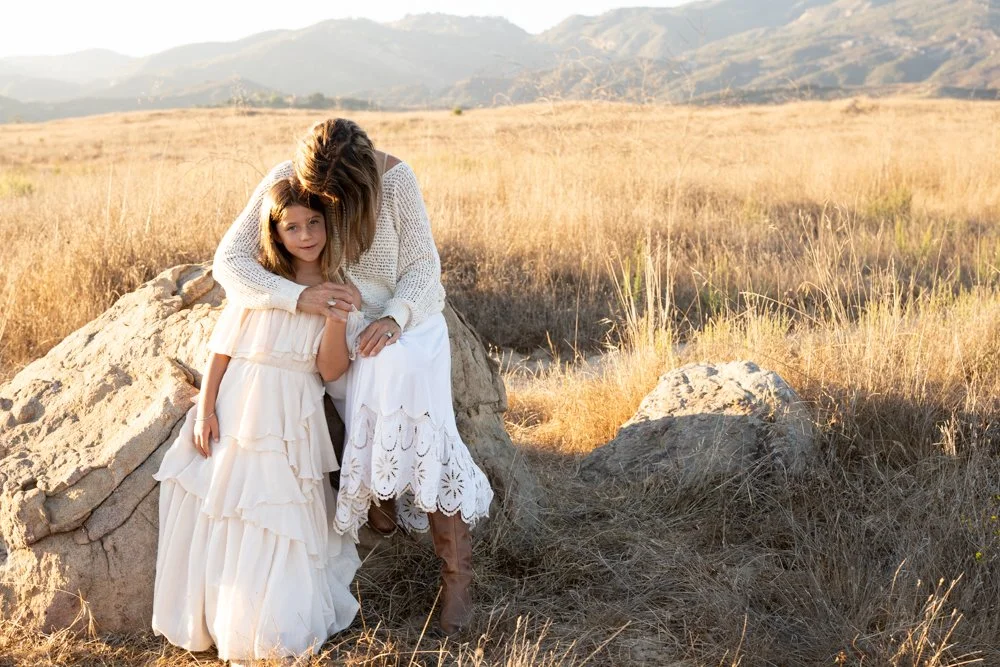 A woman and young girl sitting on a large rock in a dry, grassy field with mountains in the background, during sunset or late afternoon.