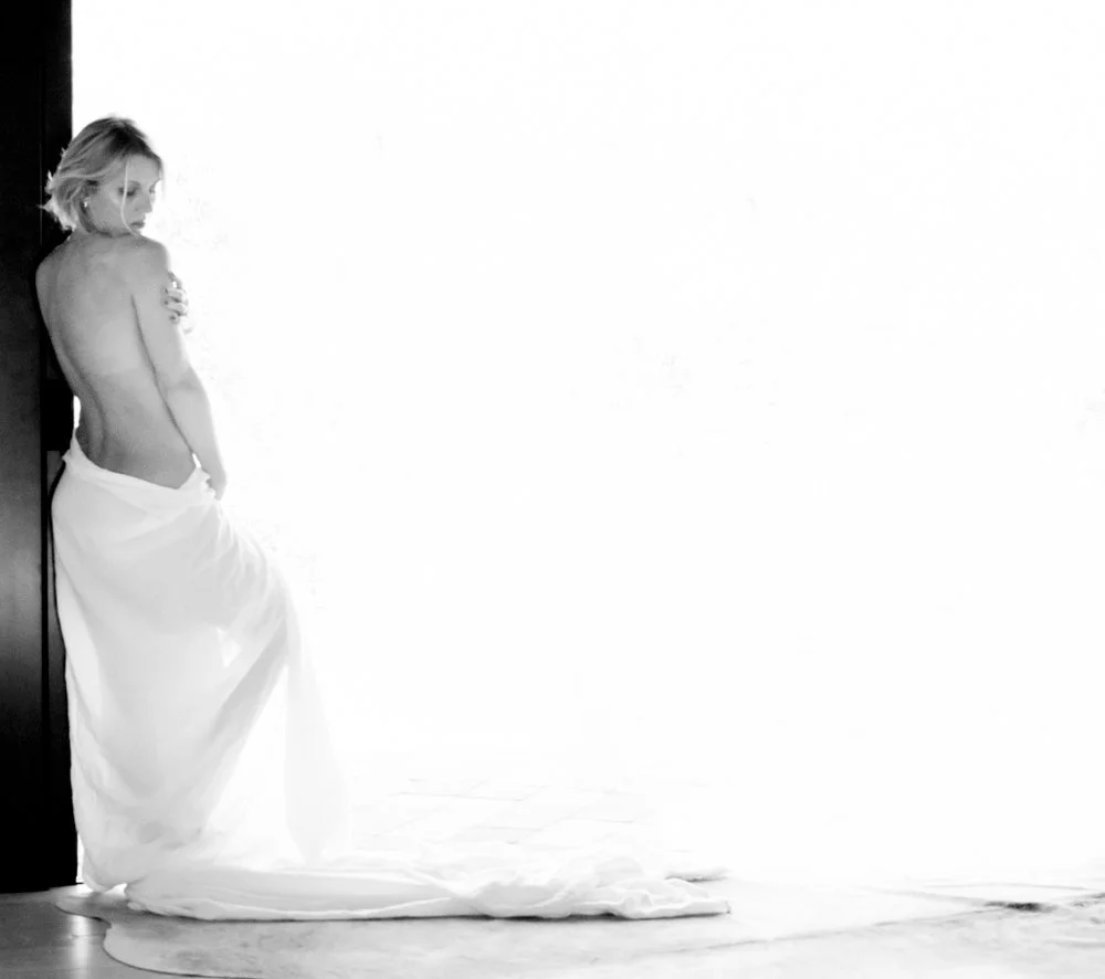 A woman with short blonde hair, standing partially covered with a white cloth, leaning against a wall in a minimalist room with a high-key white background.