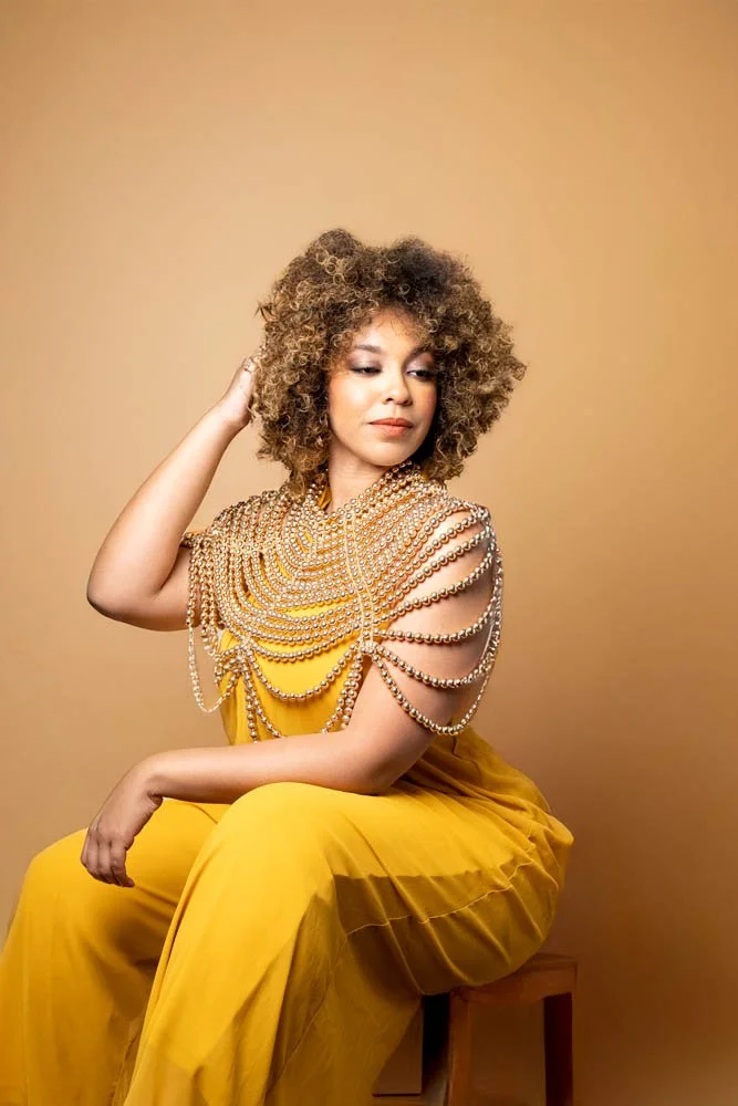 A woman with curly hair wearing a yellow dress adorned with pearl chains, sitting on a stool against a beige background.