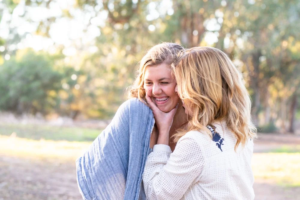 Two women sharing a joyful moment outdoors, one woman gently holds the other's face, both smiling and laughing.