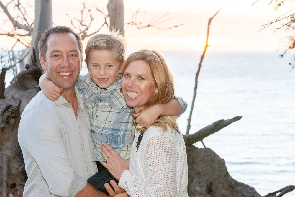 A family of three, consisting of a man, woman, and young boy, smiling and embracing outdoors near the water during sunset.